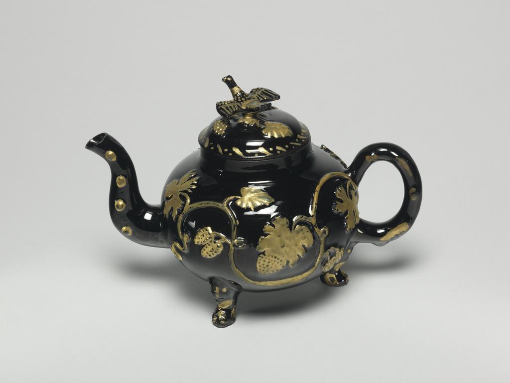 An image of Teapot and cover. Unidentified Staffordshire Factory. Dark red earthenware, the body thrown, with applied moulded handle, spout, feet, and finial, and applied moulded sprigs, the whole covered with black lead-glaze, and partly gilded. The pot has a depressed globular body supported on three lion's mask and paw feet, a crabstock handle and spout, and a domed cover with a bird finial. Both sides of the body are decorated with a coiling vine-stem with bunches of grapes and foliage in relief, which have been oil gilded. Rococo. No dimensions on Adlib (07/2014).