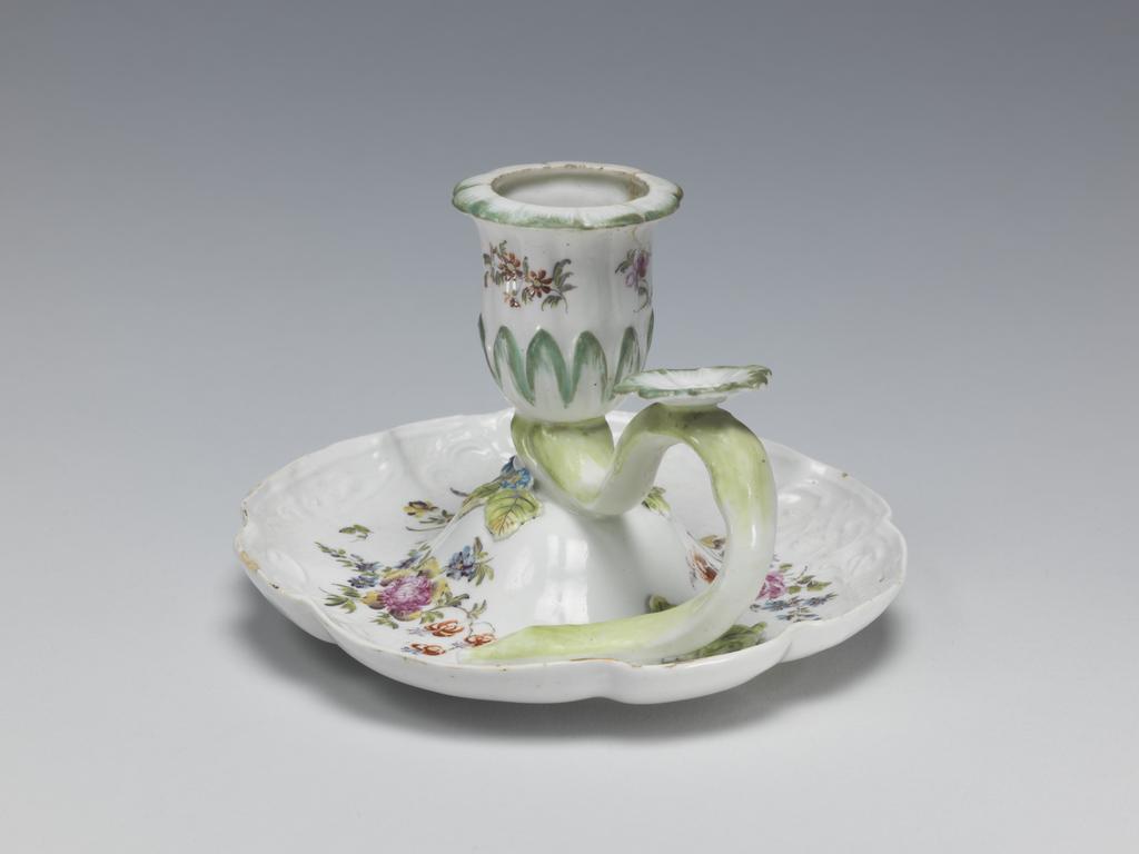 An image of Chamber Candlestick/Hand-Candlestick. Derby Porcelain Factory. The circular dish has curved sides with four large and four small lobes round the edge and a border of raised scrolls and foliage. It rises up in the centre to support the inner end of the handle and the nozzle in the form of a bell-shaped flower with a calyx of pointed leaves. The handle is in the form of a branch which curves upwards and downwards to rest on the dish. At its hightest point it has a horizontal flower-shaped thumbpiece. On the dish around the junction with the nozzle there are three blue flowers each with a pair of leaves, and another leaf lies further out beside the lower end of the handle. The dish is painted with three large sprays of mixed flowers and the nozzle with three smaller sprays of flowers of tone type. The outer edge has remnants of gilding. Soft-paste porcelain candlestick, moulded, lead-glazed, and decorated with applied flowers and leaves, and painting in polychrome enamels, height, whole, 8.2 cm, width, whole, 12.5 cm, circa 1760.