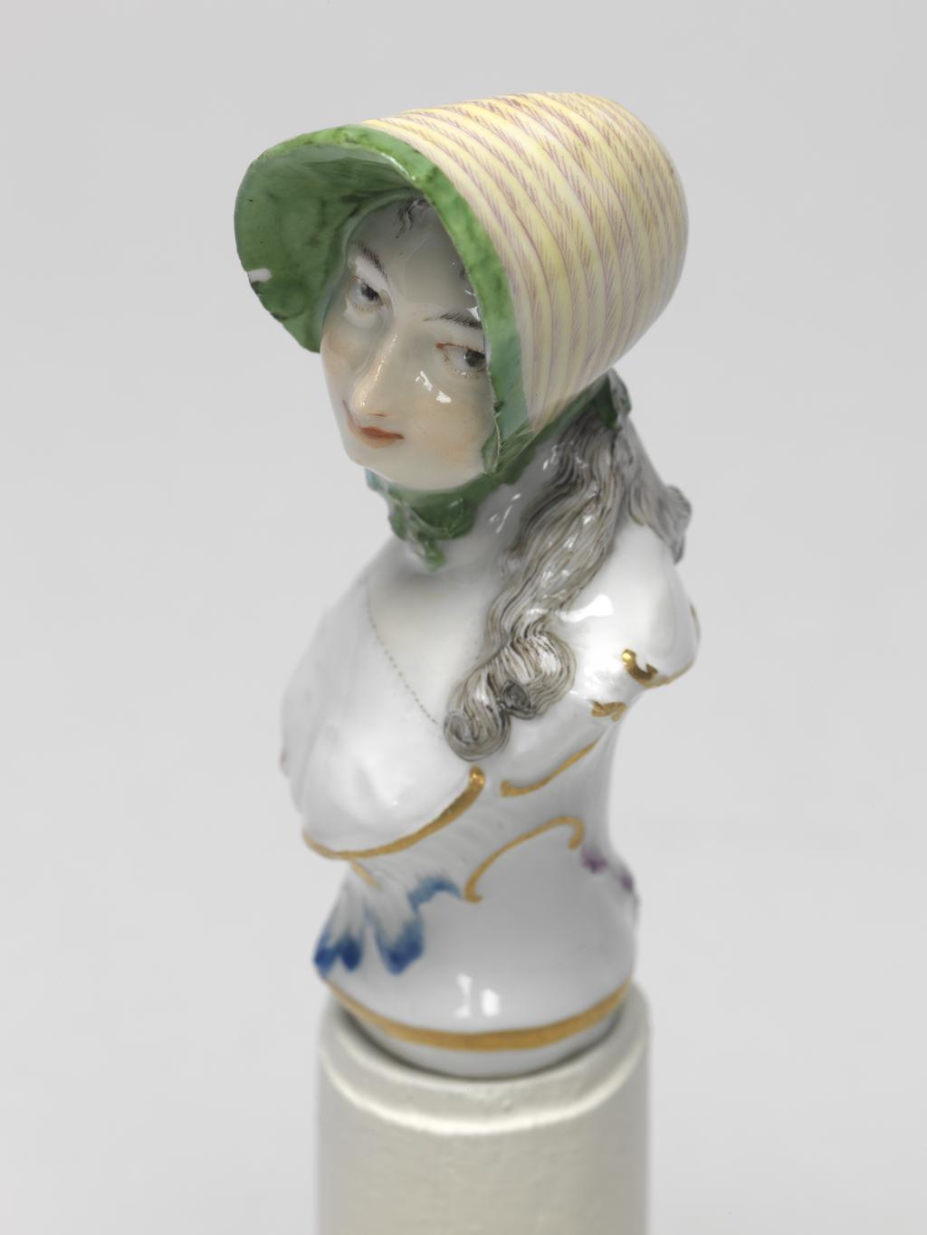 An image of Cane Handle. Nymphenburg Porcelain Factory, Bavaria. Bustelli, Franz Anton, modeller (Swiss, 1723-1763). In the form of a bust of a young woman with an integral, waisted sockle, with lightly fluted sides, and scrolls below her bust and round the back of her shoulders. The young woman's head is turned and tilted slightly to her left. She has long curling black hair which hangs down over her shouilders at the back and sides. She wears a yellow and purple striped bonnet with a green underside to the brim, and green ribbons tied under her chin, and a white bodice or chemise. The sockle is decorated with an inverted leaf-shaped outline in blue on the front, and a purple one on the back, The scrolls round its upper edge are gilded, and there is a horizontal gold band just above the lower edge. Hard-paste porcelain, press-moulded, glazed, and painted in blue, green, yellow, flesh, a little red, purple, and black enamels, and gilded; the interior is hollow and unglazed. Height, whole, 8 cm, width, shoulders, 3.5 cm, circal 1769-1760. Rococo.