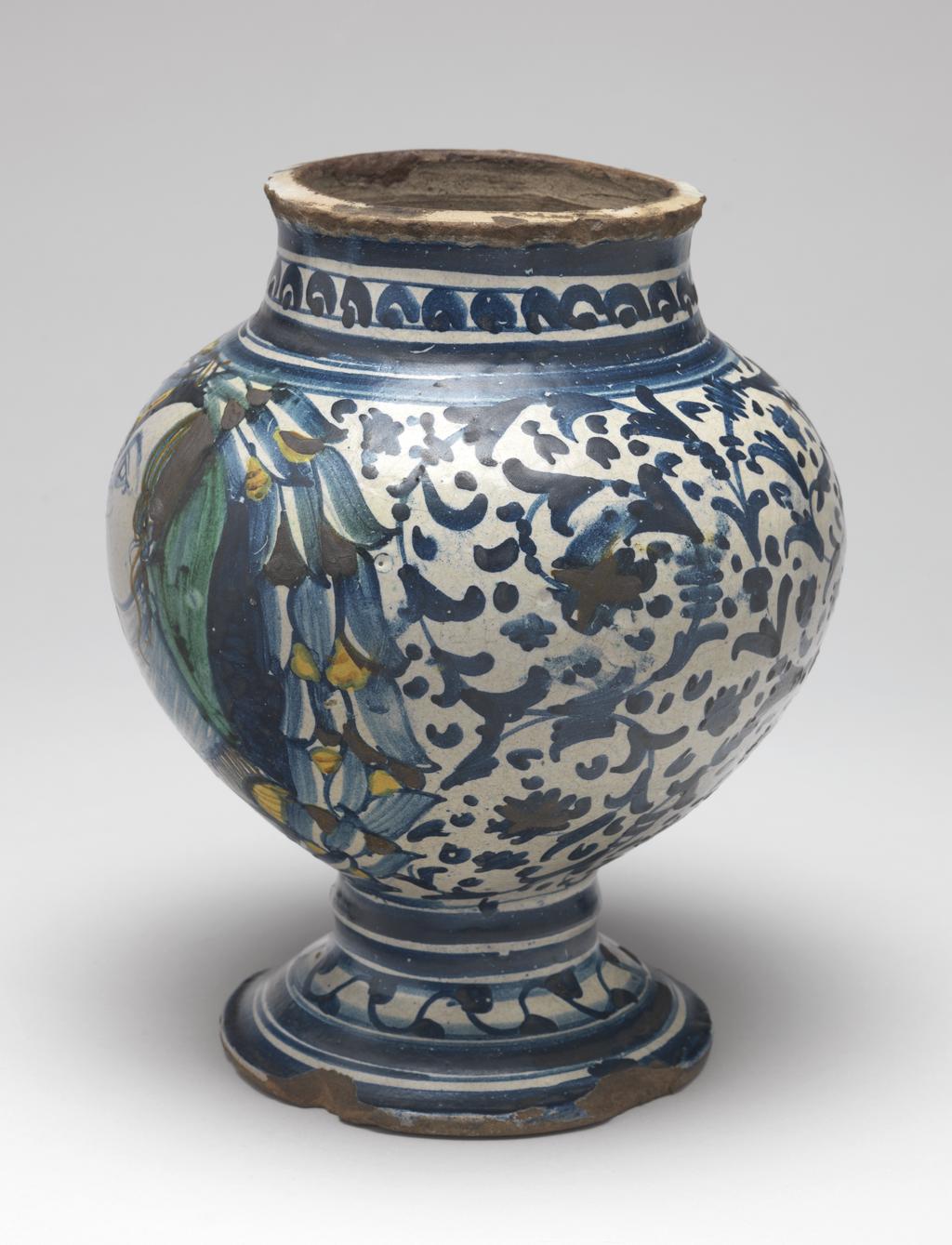 An image of Maiolica Vase. 'Master of the frowning profiles', possibly, Campania, Naples or Vietri. On the front, within a wreath of leaves and berries, is a bust of a dour young woman in profile to left facing a scroll inscribed `BEATRI' (Beatrice). The rest of the body is decorated with stylised scrolling foliage and dots. On the neck, there are semi-circles enclosing spots between horizontal bands; on the stem, two horizontal bands, and on the foot, a wavy line with spots at intervals, and concentric bands of different widths. Pinkish-buff earthenware, tin-glazed on the interior and exterior; base unglazed. Painted mainly in dark blue, with a little green, yellow, and brownish-orange. Height, whole, 21.2 cm, diameter, rim, 10.2 cm, diameter, foot, 12.0cm, diameter, wdiest part, 17.4 cm, circa 1500-1550. Renaissance. Notes: This is one of several bulbous vases and albarelli decorated with frowning profile busts, whose painter was named `Maestro dei profili corruciati' (Master of the frowning profiles) by Donatone (1993, pp. 82-3).
