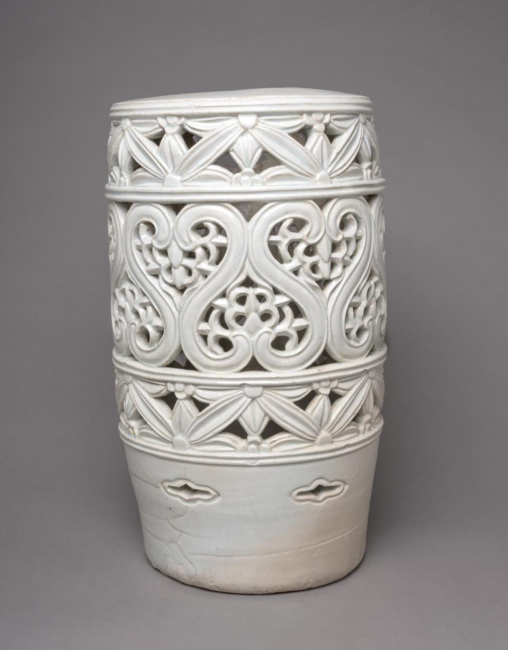 An image of Stand with floral designs. Unknown pottery, Korea. This large stand is cylindrical and has a hole in the top. The sides are divided into three horizontal zones and decorated with two bands of pierced floral designs reminiscent of Korean latticework, around a wider central pierced band with unusual S-shaped motifs enclosing leaves; above the base are small eye-shaped (ansang) openings. The glaze has a pale bluish tone. The stand has no base and the footring shows remains of coarse sand from the firing support. Porcelain, thrown, carved, pierced and glazed, height, whole, 48.7 cm, diameter, top, 26.3 cm, diameter, foot, 23.5 cm, circa 1800-1900. Choson Dynasty (1392-1910). Production Note: This piece was probably used as a stand for a flowerpot.