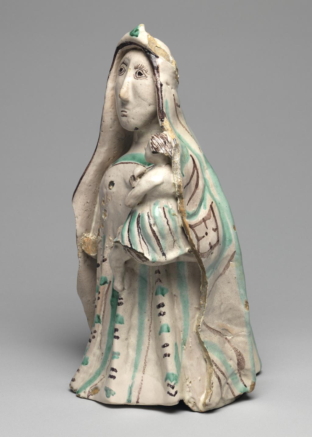 An image of Maiolica arcaica figure group. Standing Virgin and Child. Unidentified Orvieto pottery, Umbria. The Virgin stands holding the infant Christ in her left arm. Christ wears a skirt, and she wears a long pleated gown and a cloak over her head which falls down over her shoulders to the ground. The Virgin's costume is decorated with vertical rows of slanting manganese and green strokes between wide green and narrow manganese stripes. Christ's skirt has close-set stripes of alternate colours. The figure of the Virgin is hollow and the head was made separately and set into the body before firing. Greyish-buff earthenware, tin-glazed greyish-white on the exterior, and painted in manganese and copper-green, height, whole, 32.0 cm, depth, front-back, 18.0 cm, circa 1350-1450.