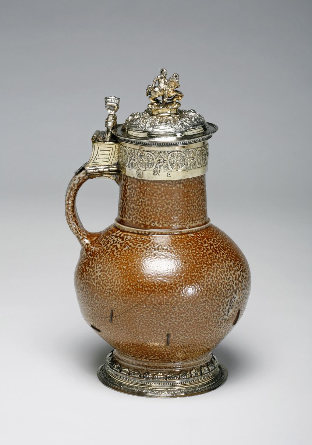 An image of Jug/Flagon. Unknown, potter, Rhineland, Frechen. Unknown, silversmith, London. The foot, neck, and top of the loop handle have silver-gilt mounts, and the thumbpiece and cover are also silver-gilt. The flat foot ring is cast and decorated with an egg and dart border. Above this is a border of dentil moulding. Cut stylised leaves hold the pottery foot in place. The neck mount is engraved with strapwork and scrolling foliage. The slightly domed cover is embossed and chased with three masks within scrolled cartouches; between are bunches of fruit on a matted ground. The urn-shaped finial stands on a gadrooned knop. The thumbpiece is a cast mask, possibly Neptune. Salt-glazed stoneware, thrown, and salt-glazed with cast, embossed, chased, and engraved silver-gilt mounts, height, to top of finial, 22.8 cm, diameter, base, 10.2 cm, width, overall, 14.4 cm, 1576-1577. Renaissance. Elizabeth I. Production Notes: Rhenish brown salt-glazed stoneware is sometimes referred to as 'tiger ware'. The body of this jug was probably made at Frechen near Cologne in the Rhineland. The silversmith's mark of an incuse leaf, has not been identified.