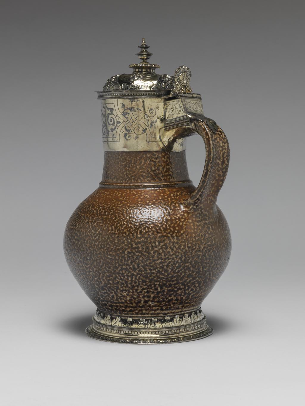 An image of Jug/Flagon. Unknown, potter, Rhineland, Frechen. Unknown, silversmith, London. The foot, neck, and top of the loop handle have silver-gilt mounts, and the thumbpiece and cover are also silver-gilt. The flat foot ring is cast and decorated with an egg and dart border. Above this is a border of dentil moulding. Cut stylised leaves hold the pottery foot in place. The neck mount is engraved with strapwork and scrolling foliage. The slightly domed cover is embossed and chased with three masks within scrolled cartouches; between are bunches of fruit on a matted ground. The urn-shaped finial stands on a gadrooned knop. The thumbpiece is a cast mask, possibly Neptune. Salt-glazed stoneware, thrown, and salt-glazed with cast, embossed, chased, and engraved silver-gilt mounts, height, to top of finial, 22.8 cm, diameter, base, 10.2 cm, width, overall, 14.4 cm, 1576-1577. Renaissance. Elizabeth I. Production Notes: Rhenish brown salt-glazed stoneware is sometimes referred to as 'tiger ware'. The body of this jug was probably made at Frechen near Cologne in the Rhineland. The silversmith's mark of an incuse leaf, has not been identified.