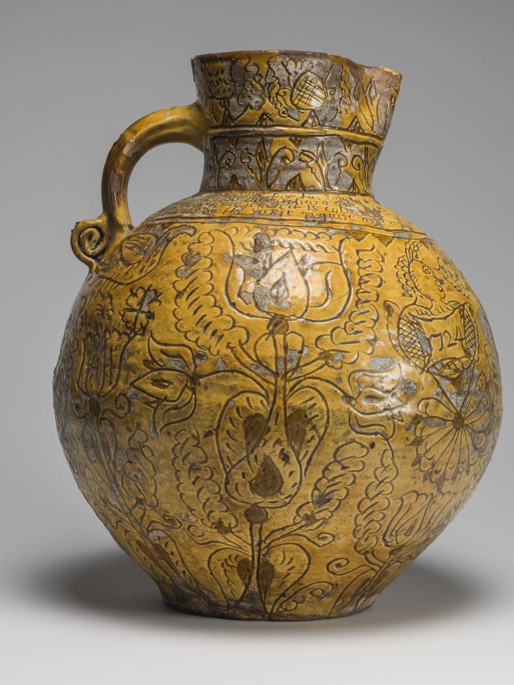 An image of Harvest Jug. Bulbous body, with a short neck pinched into a lip in front, and loop handle coiled up at lower end. On the front inscribed are the initials and date 'WH 1724', enclosed in a heart, with a thistle below and the crude busts of women, each above a cartouche with the date repeated, on either side. Below the handle is a thistle, whilst over the rest of the body except the shoulder are birds and formal tulips. On the shoulder is the inscription: 'Now i am come for to supply your workmen when in haruist dry when they do labour hard and sweat good drink is better far than meat. In winter time when it is cold i likewayes then good ale can hold All seasons do the same require and most men so strong drink desire'. Round the neck are two rows of flower motives. Earthenware, slip-coated with incised decoration under lead glaze, height, whole, 32.5 cm, dated 1724. English, North Devon.
