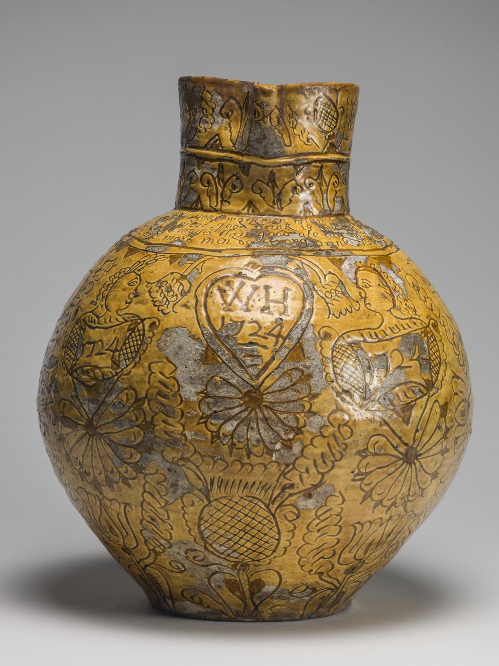 An image of Harvest Jug. Bulbous body, with a short neck pinched into a lip in front, and loop handle coiled up at lower end. On the front inscribed are the initials and date 'WH 1724', enclosed in a heart, with a thistle below and the crude busts of women, each above a cartouche with the date repeated, on either side. Below the handle is a thistle, whilst over the rest of the body except the shoulder are birds and formal tulips. On the shoulder is the inscription: 'Now i am come for to supply your workmen when in haruist dry when they do labour hard and sweat good drink is better far than meat. In winter time when it is cold i likewayes then good ale can hold All seasons do the same require and most men so strong drink desire'. Round the neck are two rows of flower motives. Earthenware, slip-coated with incised decoration under lead glaze, height, whole, 32.5 cm, dated 1724. English, North Devon.