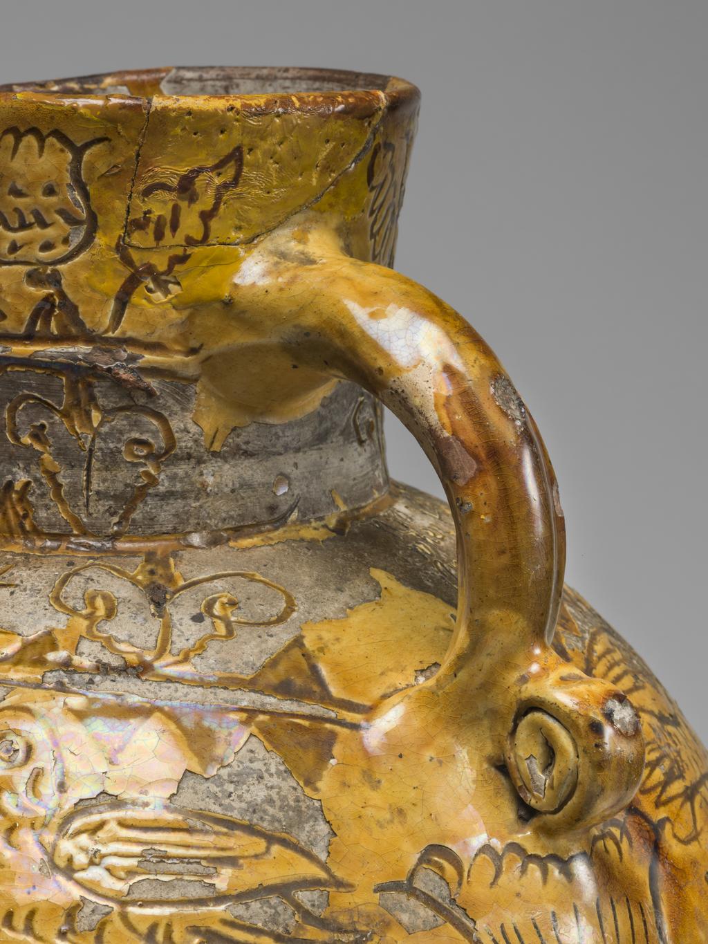 An image of Harvest Jug. Bulbous body, with a short neck pinched into a lip in front, and loop handle coiled up at lower end. On the front inscribed are the initials and date 'WH 1724', enclosed in a heart, with a thistle below and the crude busts of women, each above a cartouche with the date repeated, on either side. Below the handle is a thistle, whilst over the rest of the body except the shoulder are birds and formal tulips. On the shoulder is the inscription: 'Now i am come for to supply your workmen when in haruist dry when they do labour hard and sweat good drink is better far than meat. In winter time when it is cold i likewayes then good ale can hold All seasons do the same require and most men so strong drink desire'. Round the neck are two rows of flower motives. Earthenware, slip-coated with incised decoration under lead glaze, height, whole, 32.5 cm, dated 1724. English, North Devon.