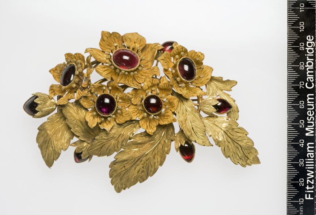 An image of Jewellery. Brooch. Carbuncles (cabochon-cut garnets), set in gold, bloomed to produce two tones and engraved. In the shape of a spray of leaves with five flowers and five buds. On the back, an oval coil of gold wire to shich the spray and the pin fastening are attached, height 8.3 cm, length 12.6 cm, circa 1840. French or English. Given by Mrs Hull Grundy.