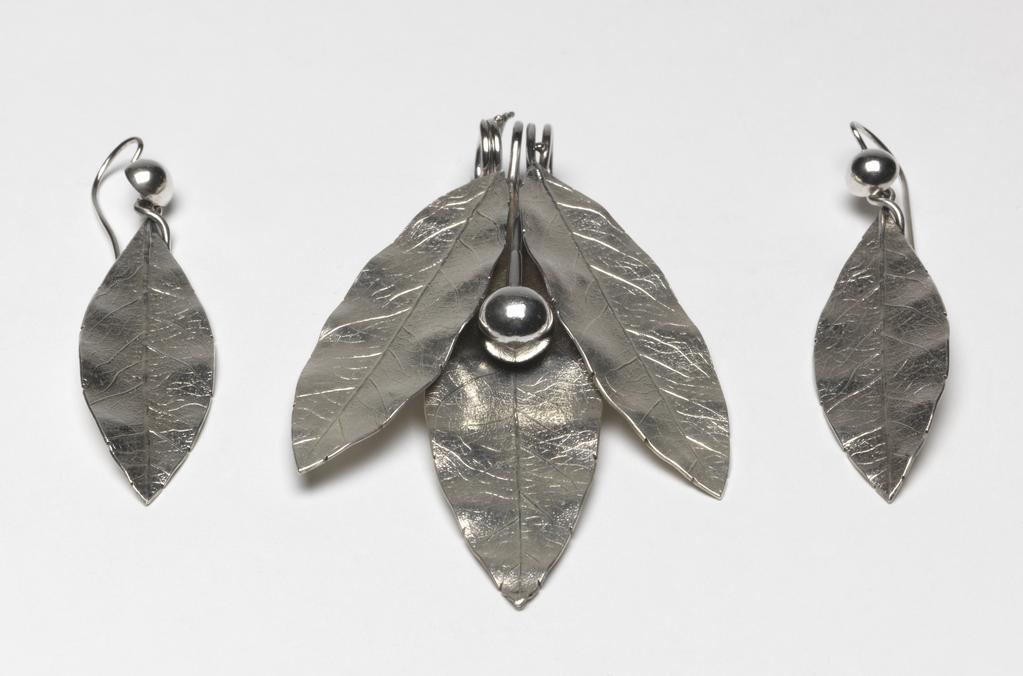 An image of Jewellery. Pendant & earrings. Phillips Brothers, London. The pendant composed of three cast laurel leaves and a berry, the stems of which are coiled to form a wide loop for suspension. Each earring is cast in the shape of a laurel leaf, the stem twisted over to form a loop into which is hooked a hemispherical berry, the stem of which is curved over and downwards to pass through a pierced ear. In original D-shaped maroon morocco case, the base lined with dark red velvet, the lid with purple silk, printed in gold below a crown 'PHILLIPS,/23, COCKSPUR ST/LONDON.' On the base of the case, a rectangular label, printed 'FENTON/33 Cranbourn St,/London' with a row of figures above in blue-black ink '3970 . . . . ' that of a later vendor. Silver, height, pendant, 8.8 cm, height, earrings, 7.2 cm, circa 1860. Victorian. Acquisition Credit: Given by Mrs J. Hull Grundy.