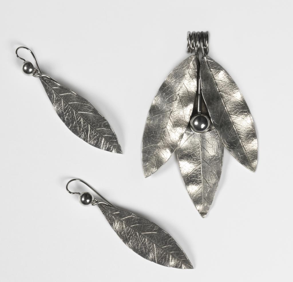 An image of Jewellery. Pendant & earrings. Phillips Brothers, London. The pendant composed of three cast laurel leaves and a berry, the stems of which are coiled to form a wide loop for suspension. Each earring is cast in the shape of a laurel leaf, the stem twisted over to form a loop into which is hooked a hemispherical berry, the stem of which is curved over and downwards to pass through a pierced ear. In original D-shaped maroon morocco case, the base lined with dark red velvet, the lid with purple silk, printed in gold below a crown 'PHILLIPS,/23, COCKSPUR ST/LONDON.' On the base of the case, a rectangular label, printed 'FENTON/33 Cranbourn St,/London' with a row of figures above in blue-black ink '3970 . . . . ' that of a later vendor. Silver, height, pendant, 8.8 cm, height, earrings, 7.2 cm, circa 1860. Victorian. Acquisition Credit: Given by Mrs J. Hull Grundy.