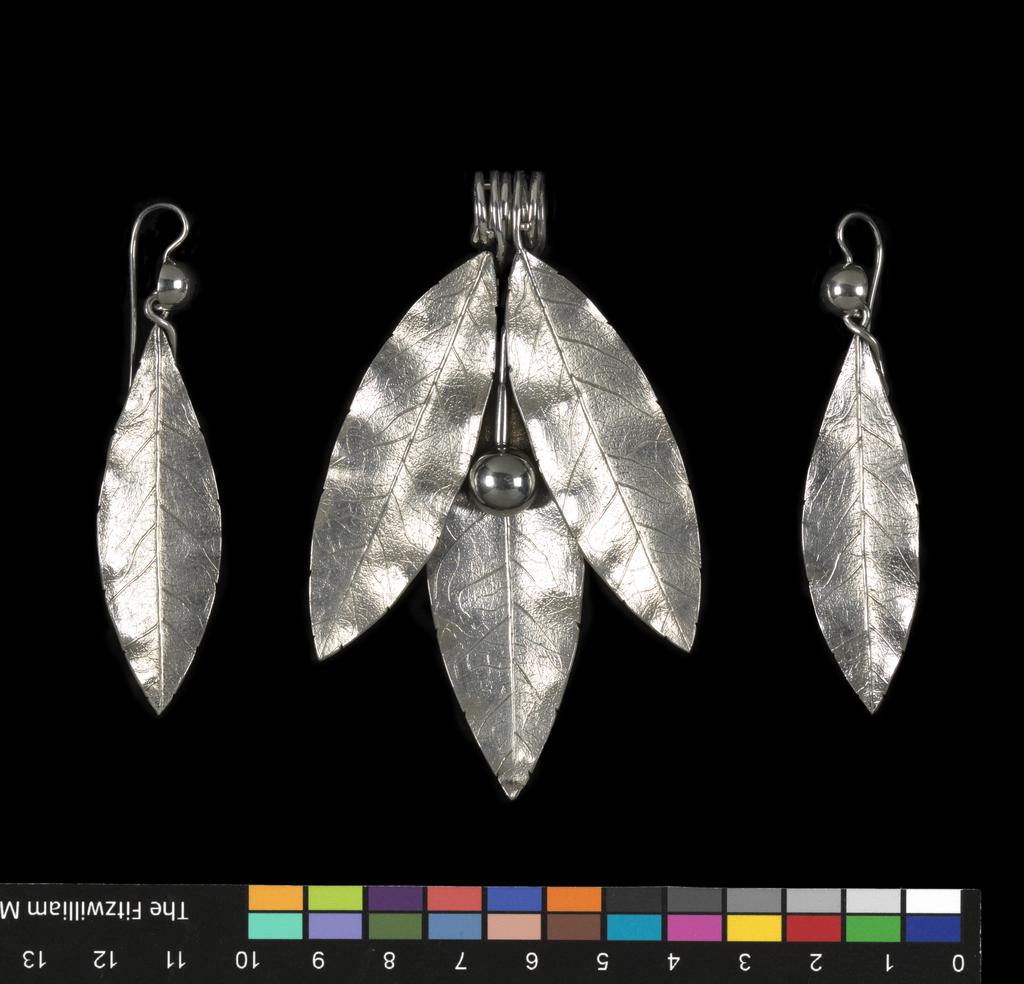 An image of Jewellery. Pendant & earrings. Phillips Brothers, London. The pendant composed of three cast laurel leaves and a berry, the stems of which are coiled to form a wide loop for suspension. Each earring is cast in the shape of a laurel leaf, the stem twisted over to form a loop into which is hooked a hemispherical berry, the stem of which is curved over and downwards to pass through a pierced ear. In original D-shaped maroon morocco case, the base lined with dark red velvet, the lid with purple silk, printed in gold below a crown 'PHILLIPS,/23, COCKSPUR ST/LONDON.' On the base of the case, a rectangular label, printed 'FENTON/33 Cranbourn St,/London' with a row of figures above in blue-black ink '3970 . . . . ' that of a later vendor. Silver, height, pendant, 8.8 cm, height, earrings, 7.2 cm, circa 1860. Victorian. Acquisition Credit: Given by Mrs J. Hull Grundy.