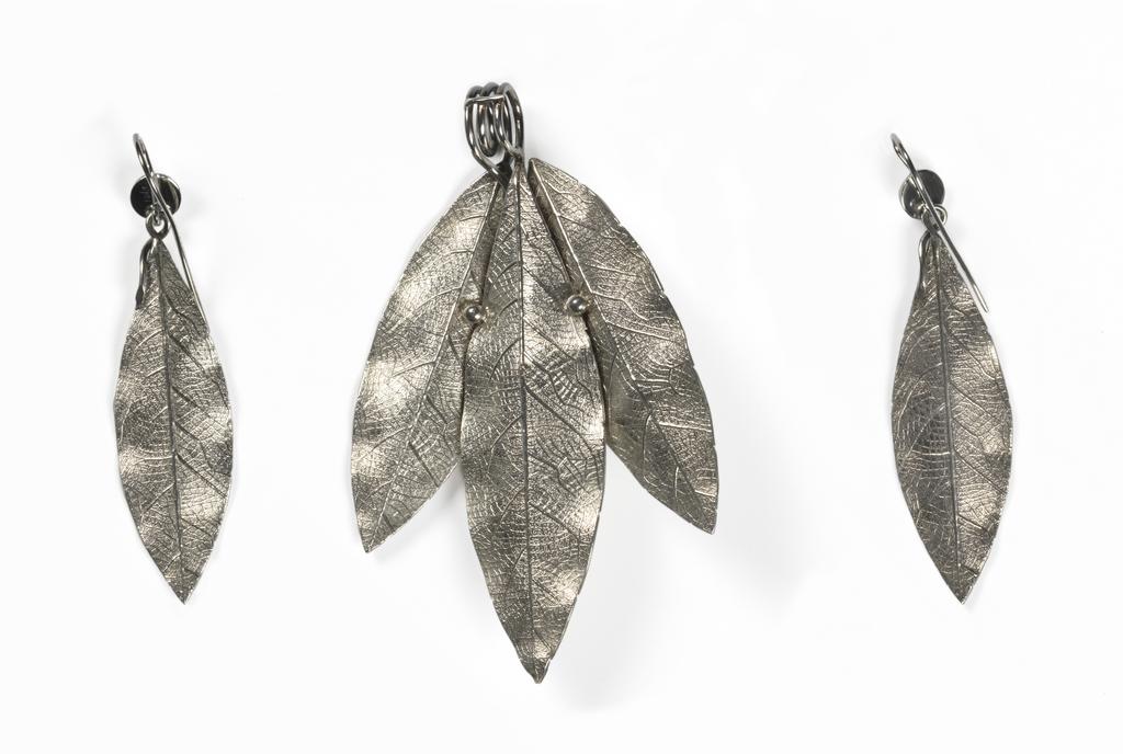 An image of Jewellery. Pendant & earrings. Phillips Brothers, London. The pendant composed of three cast laurel leaves and a berry, the stems of which are coiled to form a wide loop for suspension. Each earring is cast in the shape of a laurel leaf, the stem twisted over to form a loop into which is hooked a hemispherical berry, the stem of which is curved over and downwards to pass through a pierced ear. In original D-shaped maroon morocco case, the base lined with dark red velvet, the lid with purple silk, printed in gold below a crown 'PHILLIPS,/23, COCKSPUR ST/LONDON.' On the base of the case, a rectangular label, printed 'FENTON/33 Cranbourn St,/London' with a row of figures above in blue-black ink '3970 . . . . ' that of a later vendor. Silver, height, pendant, 8.8 cm, height, earrings, 7.2 cm, circa 1860. Victorian. Acquisition Credit: Given by Mrs J. Hull Grundy.
