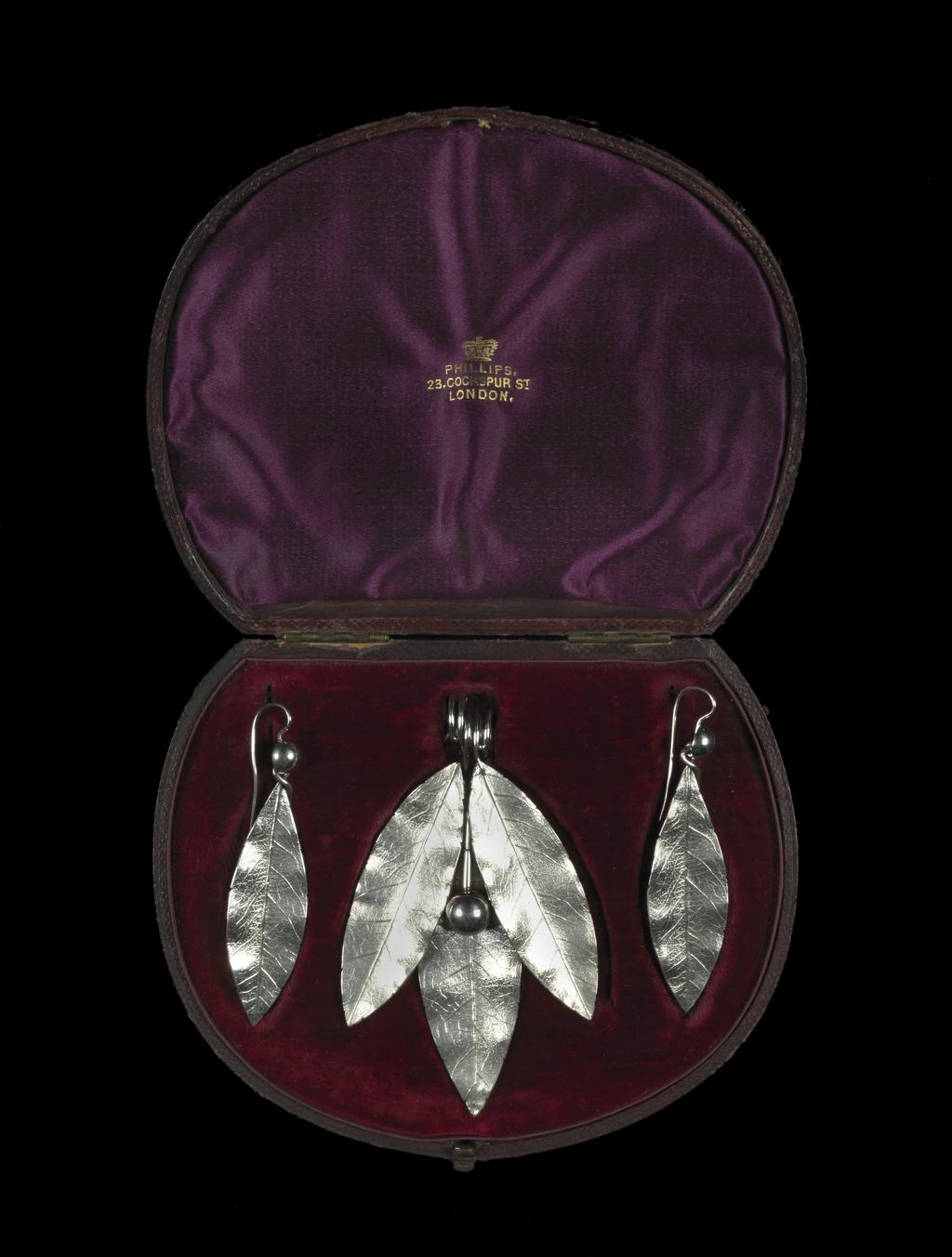 An image of Jewellery. Pendant & earrings. Phillips Brothers, London. The pendant composed of three cast laurel leaves and a berry, the stems of which are coiled to form a wide loop for suspension. Each earring is cast in the shape of a laurel leaf, the stem twisted over to form a loop into which is hooked a hemispherical berry, the stem of which is curved over and downwards to pass through a pierced ear. In original D-shaped maroon morocco case, the base lined with dark red velvet, the lid with purple silk, printed in gold below a crown 'PHILLIPS,/23, COCKSPUR ST/LONDON.' On the base of the case, a rectangular label, printed 'FENTON/33 Cranbourn St,/London' with a row of figures above in blue-black ink '3970 . . . . ' that of a later vendor. Silver, height, pendant, 8.8 cm, height, earrings, 7.2 cm, circa 1860. Victorian. Acquisition Credit: Given by Mrs J. Hull Grundy.