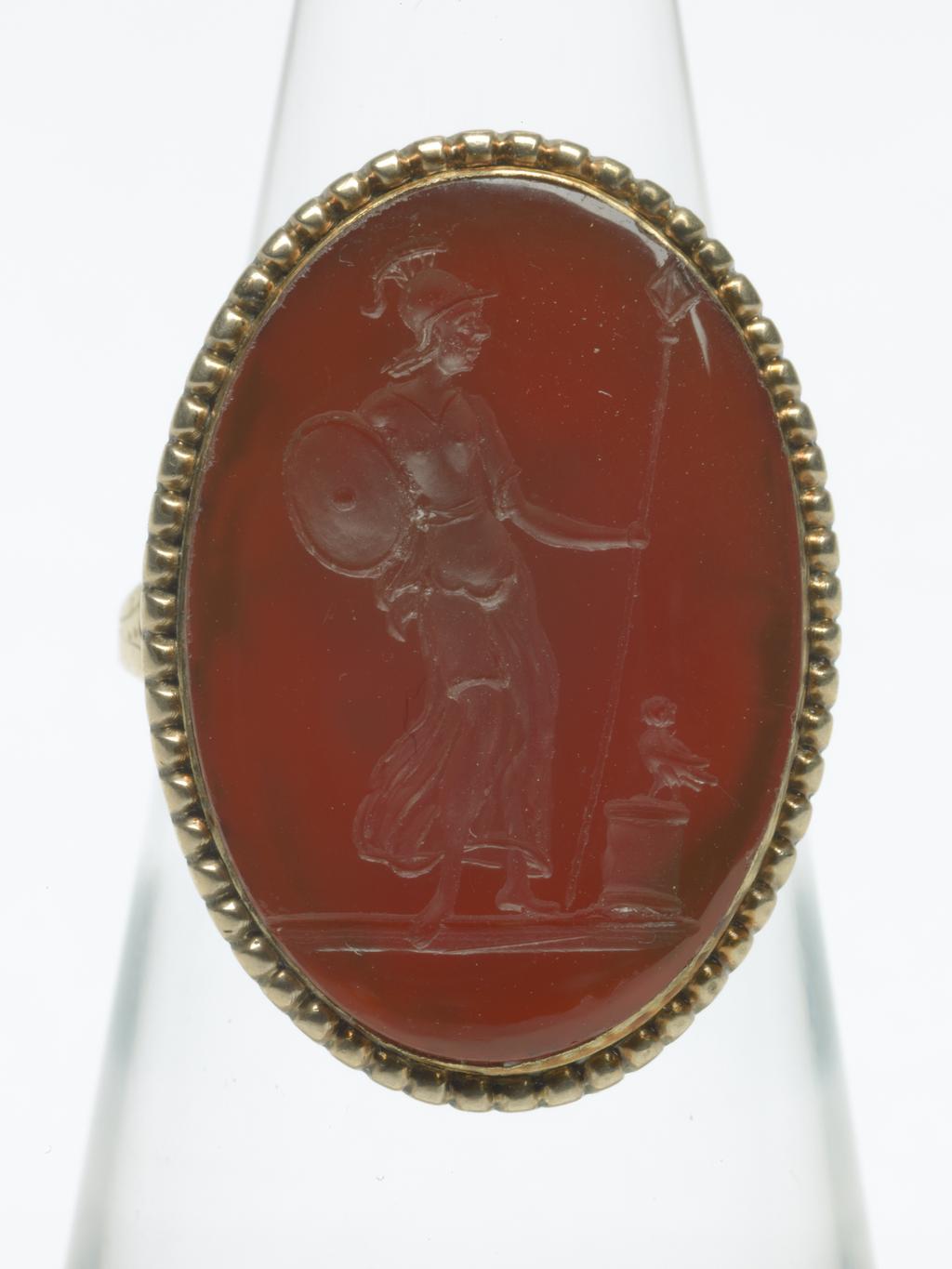 An image of Jewellery. Ring. Intaglio seal. Unknown maker, probably English. Red carnelian intaglio depicting standing figure of Minerva, wearing plumed helmet and chiton, leaning on a spear and carrying her shield on her arm, head turned towards the left, owl on a low altar beside her. Pearled border and ground line. The device also appears on the reverse of a medallic portrait of Gustavus III of Sweden. Gold with pointed oval bezel, beaded border and closed back; square hoop expanding at the shoulders, ornamented with bright cutting. Gold ring, red carnelian intaglio, height, intaglio, 22 mm, width, intaglio, 14 mm, circa 1700-1800.