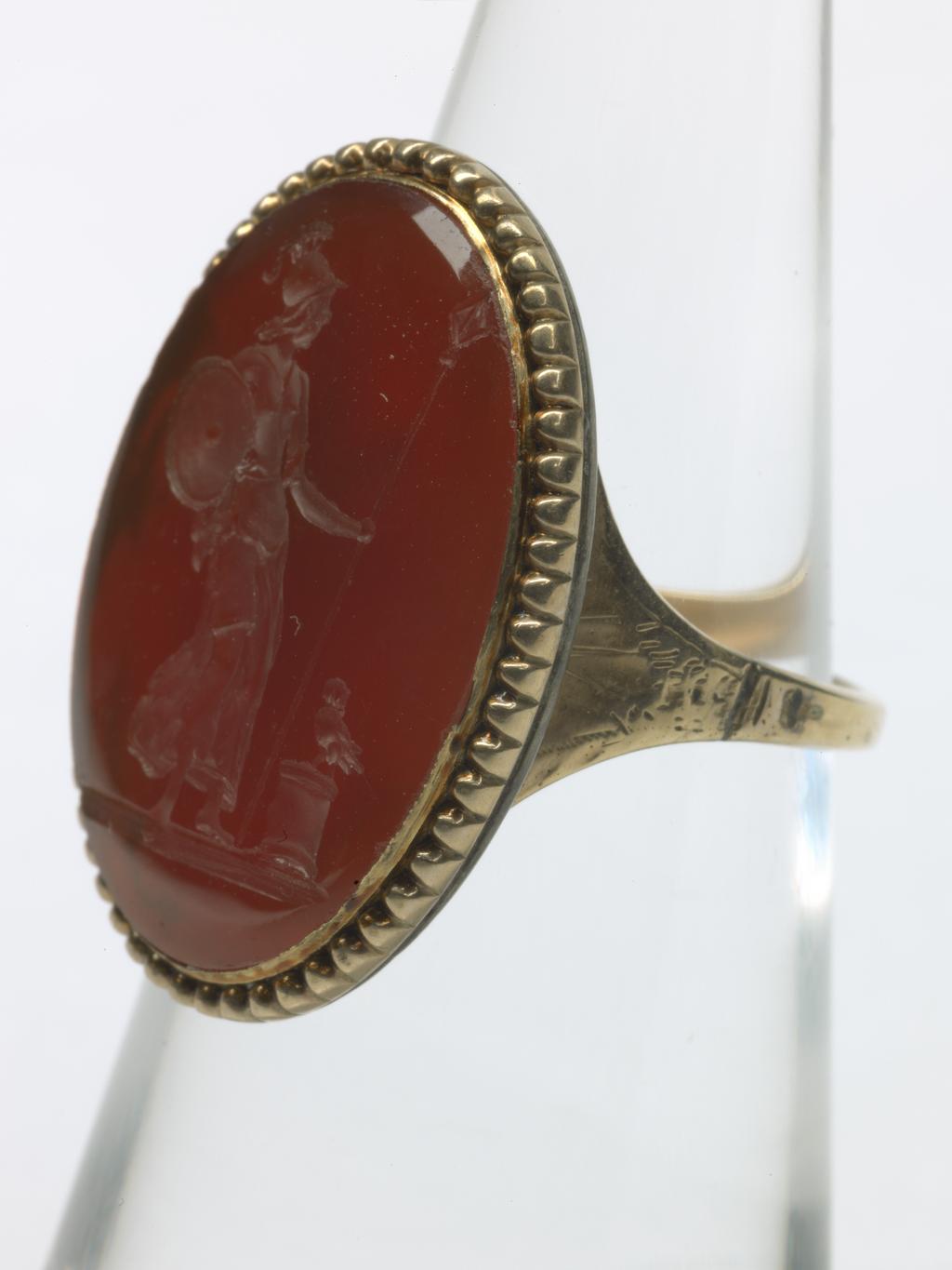 An image of Jewellery. Ring. Intaglio seal. Unknown maker, probably English. Red carnelian intaglio depicting standing figure of Minerva, wearing plumed helmet and chiton, leaning on a spear and carrying her shield on her arm, head turned towards the left, owl on a low altar beside her. Pearled border and ground line. The device also appears on the reverse of a medallic portrait of Gustavus III of Sweden. Gold with pointed oval bezel, beaded border and closed back; square hoop expanding at the shoulders, ornamented with bright cutting. Gold ring, red carnelian intaglio, height, intaglio, 22 mm, width, intaglio, 14 mm, circa 1700-1800.