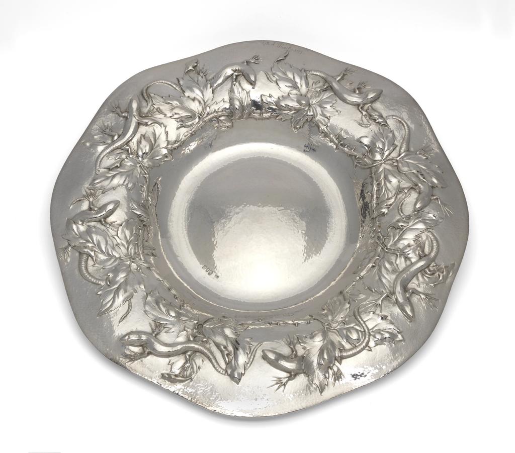 An image of Silver dish. Marks, Gilbert Leigh (British, 1861-1905). The dish was raised and the lizards and foliage embossed and chased. The lizards have a small piece of silver seamed under the jaw and neck, which allows the head to stand proud of the dish. The background to the dish is spot hammered. Height to rim 5 cm (variable), diameter (across rim) 42 cm, 1898-1899. Arts and Crafts movement.