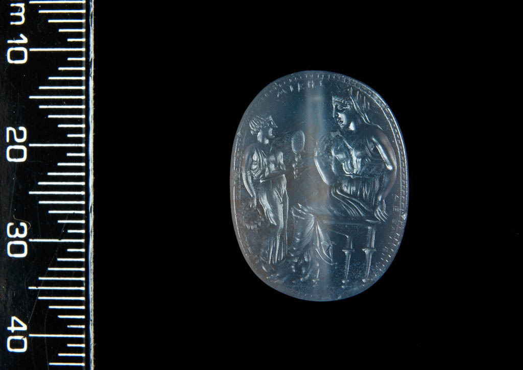An image of Engraved gems/scaraboid. Seated woman in sleeveless chiton, profile to right, looking in mirror held by servant girl. Inscription 'belonging to Mike' and 'made by Dexamenos'. Intaglio cutting, sapphirine chalcedony, height 22mm, width 17mm, depth 8mm. Classical Greek Period, circa 450-425 B.C.