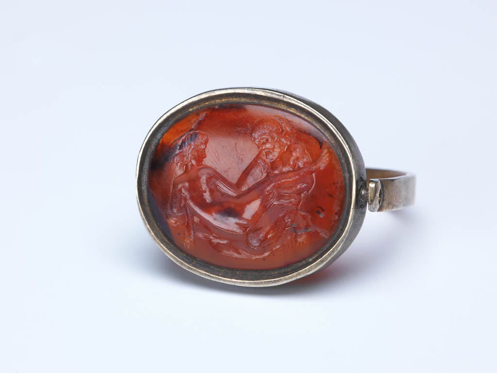 An image of Engraved gems/sealstone/intaglio. A love-scene, with Hercules and Omphale on a bed covered with the lion-skin. Mount: silver swivel-ring, type 3. Intaglio cutting, sard, height 19 mm, width 15.5 mm, circa 1500- circa 1700.
