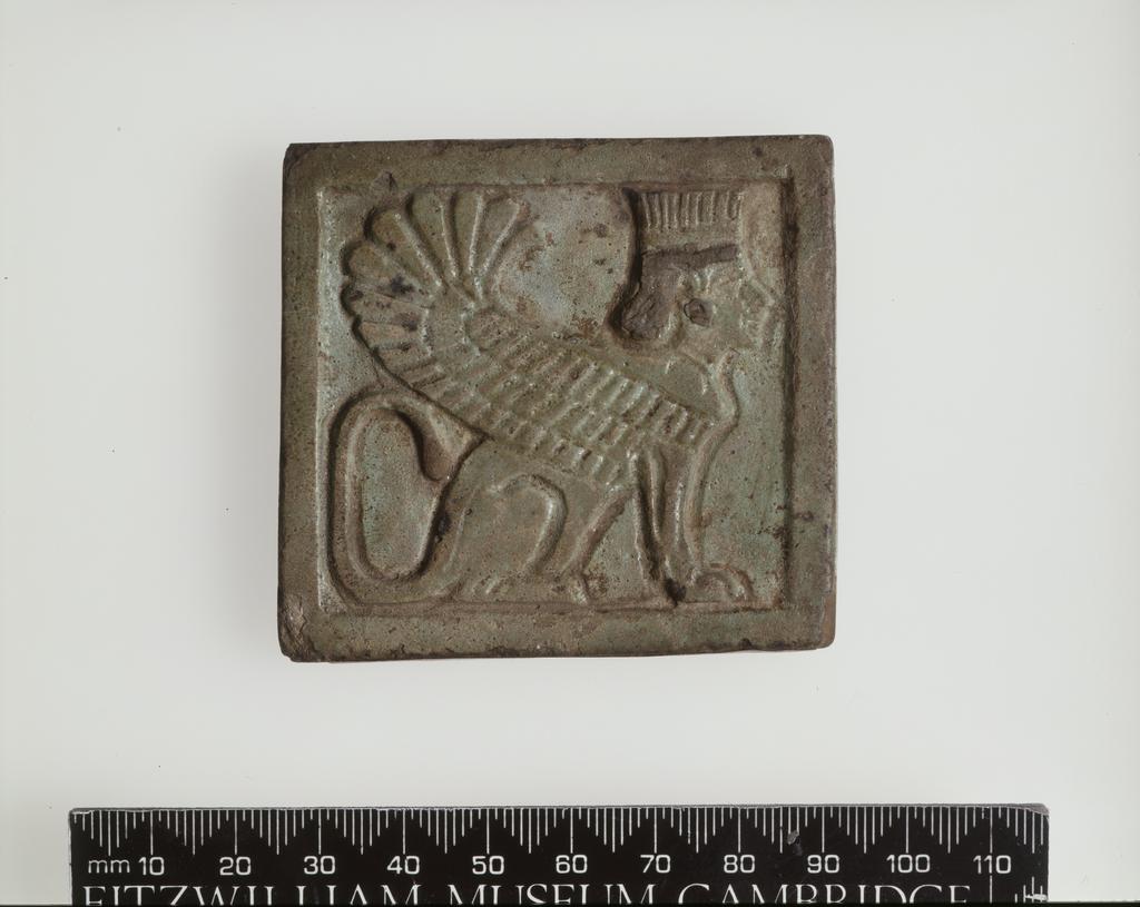 An image of Plaque, with winged Persian sphinx, pierced for suspension. Find Spot/Production Place: Egypt. Faience, length 6.2 cm, width 6.6 cm, circa 525-404 B.C. Twenty-seventh Dynasty.