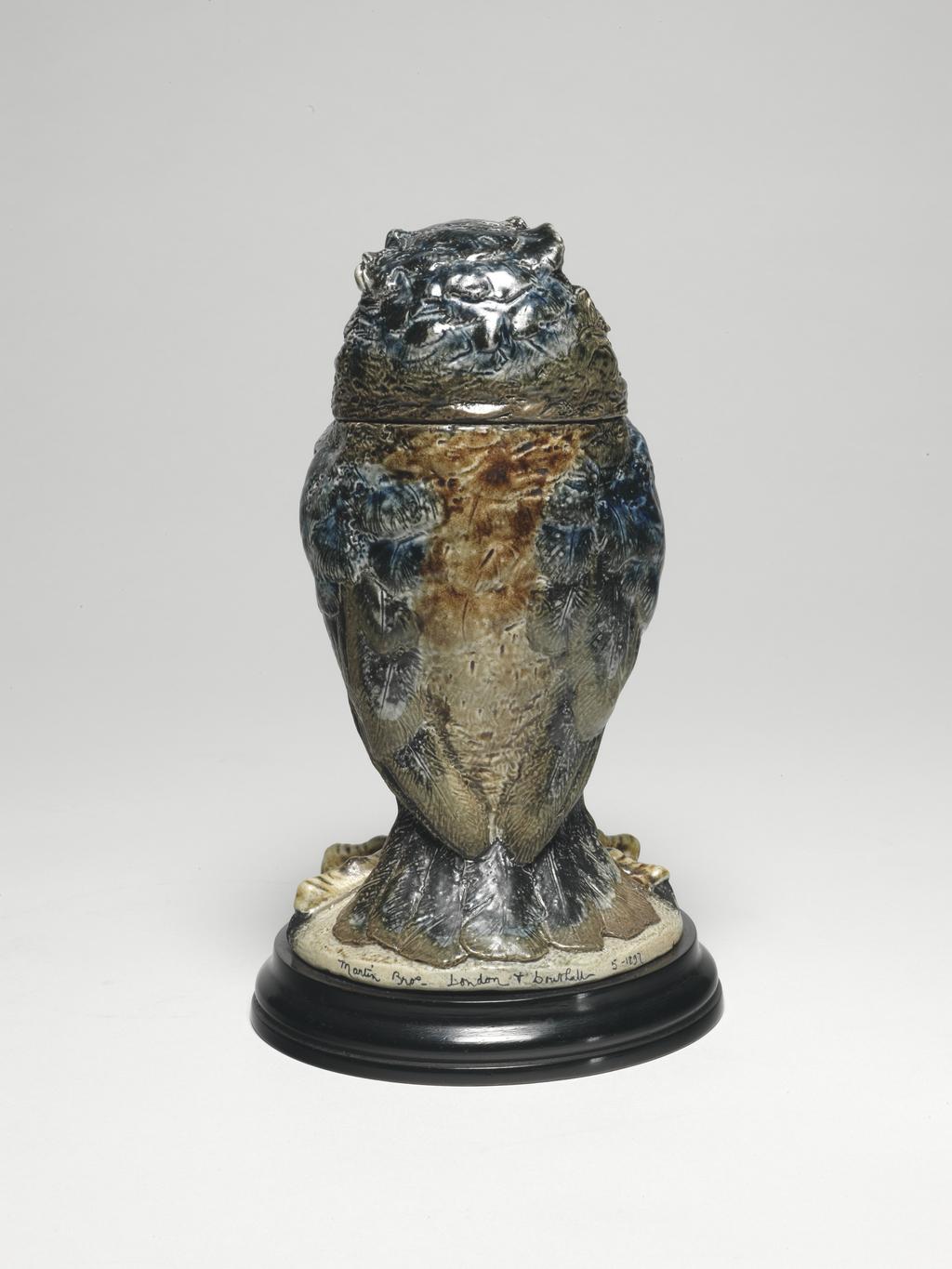 An image of Wally Bird. Martin Brothers. Stoneware tobacco jar and cover, hand-modelled onto a press-moulded body into the shape of a grotesque bird, painted in cobalt blue and brown, and salt-glazed; the base fixed with nails onto a turned, circular wooden base-plate. Height (whole) 23.2 cm. Dated 1897.