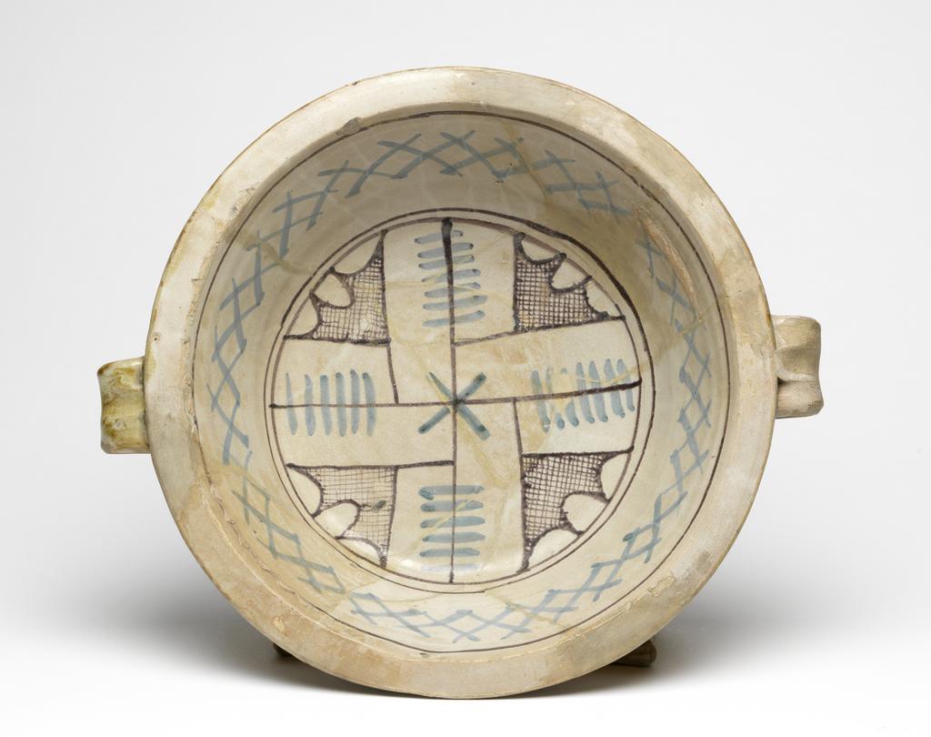 An image of Maiolica arcaica blu. Two-handled bowl. Unidentified pottery, probably Umbria, Orvieto or Tuscany. Shape 39. Circular with flat base, sloping sides, narrow flat rim and two wide strap handles with a thumb print at the top. Inside, within two manganese concentric circles, there is a double-armed cross with cross-hatching and three scalloped lines between the arms; on the sides, blue criss-crosses and a manganese band. Pale buff earthenware. The exterior and base are lead-glazed pale brown; the interior is tin-glazed pale grey. Painted in manganese and blue. Height, whole, 9.7 cm, diameter, base, 18.8 cm, width, whole, 34.0 cm, circa 1350-1425. Late Medieval.