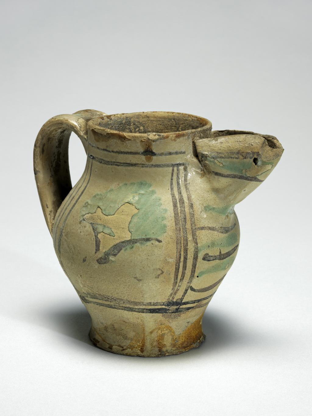 An image of Tin-glazed earthenware jug. Maiolica arcaica. Painted in green and brown with panels, leaves and stripes. Cream earthenware, remnants of lead glaze on lower part and interior, the rest tin-glazed, painted in green and manganese-brown, height 10.3cm, diameter (base) 5.5cm, width (body) 8.8cm, width (handle to spout) 11.4cm, circa 1250-1300. From Umbria, Italy. Medieval.
