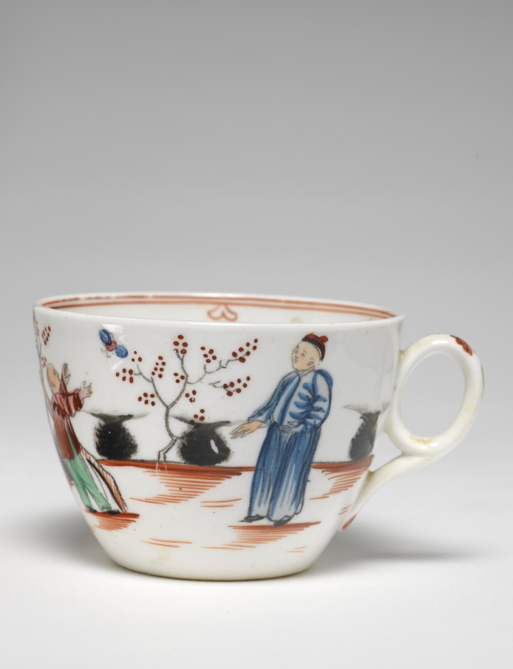 An image of Cup and saucer. New Hall Porcelain Factory, Staffordshire. Bute shape cup with oval ring handle. Circular saucer with curved sides, standing on a footring. The interior of the cup is decorated at the bottom with a row of red grasses, and round the rim with two narrow red lines under which are four inverted hearts with a circle below. The exterior is decorated with a continuous Chinese scene: two fuzzy black bushes behind a red fence; a woman wearing green and puce clothing, stands holding a sceptre; a spiky black bush and a flowering tree; a boy dressed in green and dark puce who is trying to catch a butterfly; two black bushes flanking a flowering tree; a standing man dressed in blue, and another black bush. The ground is indicated by horizontal red strokes. On the back of the handle in puce are five dots, a stylized leaf, a vertical stroke, and at the lower end two leaves. The saucer is decorated inside with a Chinese scene with spiky black rocks, a flowering shrub and flowering tree; a boy holding up arms to a butterfly, and a man and woman standing side by side, dressed similarly to those on the cup. The border is similar to that on the cup with six heart motifs. Pattern 421. Saucer of hard-paste porcelain, press-moulded, painted overglaze in enamels; cup is possibly bone china, lead-glazed, height, cup, 6 cm, width, cup, 10.5 cm, diameter, cup, 8.5 cm, height, saucer, 3 cm, diameter, saucer, 14.3 cm, circa 1800- 1805. Chinese Style.