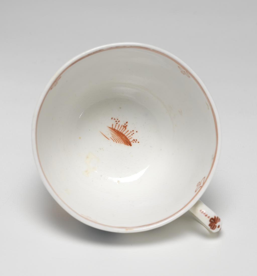 An image of Cup and saucer. New Hall Porcelain Factory, Staffordshire. Bute shape cup with oval ring handle. Circular saucer with curved sides, standing on a footring. The interior of the cup is decorated at the bottom with a row of red grasses, and round the rim with two narrow red lines under which are four inverted hearts with a circle below. The exterior is decorated with a continuous Chinese scene: two fuzzy black bushes behind a red fence; a woman wearing green and puce clothing, stands holding a sceptre; a spiky black bush and a flowering tree; a boy dressed in green and dark puce who is trying to catch a butterfly; two black bushes flanking a flowering tree; a standing man dressed in blue, and another black bush. The ground is indicated by horizontal red strokes. On the back of the handle in puce are five dots, a stylized leaf, a vertical stroke, and at the lower end two leaves. The saucer is decorated inside with a Chinese scene with spiky black rocks, a flowering shrub and flowering tree; a boy holding up arms to a butterfly, and a man and woman standing side by side, dressed similarly to those on the cup. The border is similar to that on the cup with six heart motifs. Pattern 421. Saucer of hard-paste porcelain, press-moulded, painted overglaze in enamels; cup is possibly bone china, lead-glazed, height, cup, 6 cm, width, cup, 10.5 cm, diameter, cup, 8.5 cm, height, saucer, 3 cm, diameter, saucer, 14.3 cm, circa 1800- 1805. Chinese Style.
