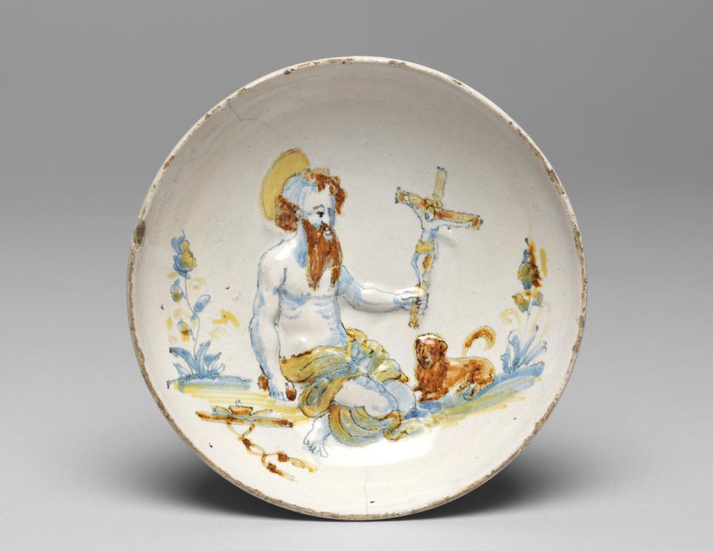 An image of Maiolica Bowl. St Jerome in the Wilderness. Unknown potter, Umbria, Deruta. Shape 76. Circular with curved sides, standing on a low footring, pierced by a hole incorrectly placed for suspension the right way up. The Saint kneels facing towards the right, holding a crucifix in his left hand and a stone in his right. A lion crouches on his left and a cardinal's hat and tassels lie on the ground to his right. In the background on either side there is a plant. Light greyish-buff earthenware. Moulded with the design in relief, and tin-glazed overall; unevenly and thinly on the reverse. Painted in blue, yellow, and brownish-orange, height, whole, 4.4 cm, diameter, whole, 15.2 cm, circa 1575-1625. Renaissance. Notes(s): St Jerome was a common subject on lustred maiolica in the first half of the sixteenth century. St Jerome (c. 341-420) was one of the four Latin fathers of the early Church, whose greatest achievement was the production of a Latin text of the Bible, later known as the Vulgate. On this bowl, as on most Deruta and Gubbio lustreware, he is shown as a hermit, kneeling in penitence in the Syrian desert where he spent five years before he was ordained. The lion became his companion after he had taken a thorn from its paw, much later in his life when he was a monk at Bethlehem. This bowl is painted in the compendiario style, which was adopted in Deruta during the last third of the sixteenth century and continued for much of the seventeenth.