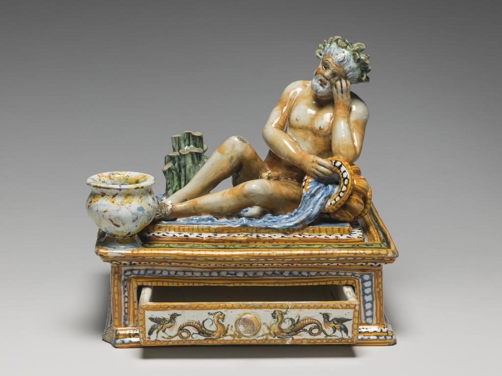 An image of Maiolica. Inkstand with drawer. Patanazzi family, workshop, probably (Italian, 16th-17th cs), The Marches, Urbino. Rectangular with projecting mouldings at the top and bottom, and a drawer at the front. A slightly raised platform on the top supports a river god who reclines with his left elbow resting on an urn with water flowing from it. Behind him to the left there is a clump of reeds and on the front left corner, a vase for ink. The river god, reeds and water are coloured naturalistically. The sides are decorated with grotesques in narrow panels framed by yellow and orange, and blue and white, borders. The inkwell is spattered to resemble coloured marble, and the urn is streaked in yellow and orange. Earthenware, tin-glazed overall except for the edge of the base. Painted in blue, green, yellow, orange, manganese-purple, black, and white, height, whole, 21.0 cm, width, whole, 22.1 cm, depth, whole, 17.8 cm, circa 1575-1605. Renaissance.