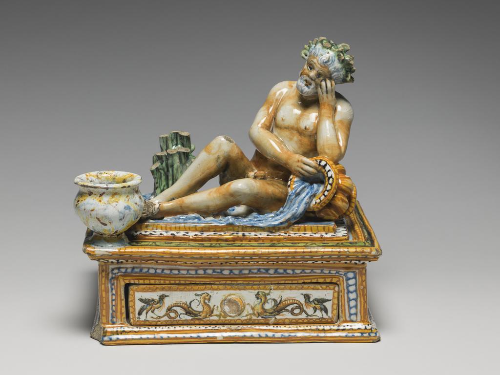 An image of Maiolica. Inkstand with drawer. Patanazzi family, workshop, probably (Italian, 16th-17th cs), The Marches, Urbino. Rectangular with projecting mouldings at the top and bottom, and a drawer at the front. A slightly raised platform on the top supports a river god who reclines with his left elbow resting on an urn with water flowing from it. Behind him to the left there is a clump of reeds and on the front left corner, a vase for ink. The river god, reeds and water are coloured naturalistically. The sides are decorated with grotesques in narrow panels framed by yellow and orange, and blue and white, borders. The inkwell is spattered to resemble coloured marble, and the urn is streaked in yellow and orange. Earthenware, tin-glazed overall except for the edge of the base. Painted in blue, green, yellow, orange, manganese-purple, black, and white, height, whole, 21.0 cm, width, whole, 22.1 cm, depth, whole, 17.8 cm, circa 1575-1605. Renaissance.