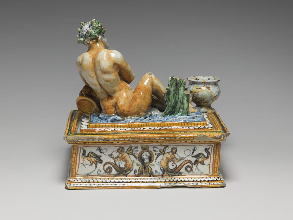 An image of Maiolica. Inkstand with drawer. Patanazzi family, workshop, probably (Italian, 16th-17th cs), The Marches, Urbino. Rectangular with projecting mouldings at the top and bottom, and a drawer at the front. A slightly raised platform on the top supports a river god who reclines with his left elbow resting on an urn with water flowing from it. Behind him to the left there is a clump of reeds and on the front left corner, a vase for ink. The river god, reeds and water are coloured naturalistically. The sides are decorated with grotesques in narrow panels framed by yellow and orange, and blue and white, borders. The inkwell is spattered to resemble coloured marble, and the urn is streaked in yellow and orange. Earthenware, tin-glazed overall except for the edge of the base. Painted in blue, green, yellow, orange, manganese-purple, black, and white, height, whole, 21.0 cm, width, whole, 22.1 cm, depth, whole, 17.8 cm, circa 1575-1605. Renaissance.