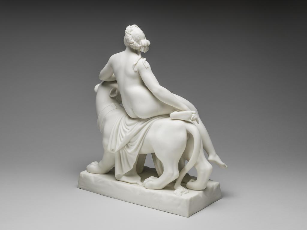An image of Ariadne on the Panther or Voluptuousness. Minton Factory, Stoke on Trent. After the sculpture by John Heinrich von Dannecker. The figures are supported on a rectangular base with slightly sloping sides and indentations suggesting it is rocky. The panther is walking to the right with its left fore and hind legs forward, its tail between its hind feet, and its head turned towards the viewer, baring its fangs. The nude Ariadne is seated on its back with her right leg extended and her left bent.  Her head is in profile, and she rests her left fore arm on the panther's head. With her left hand she holds up a long drape which extends down to the base at the front, over the panther's back beneath her legs, and down to the back of the base. Creamy-white parian soft-paste porcelain, slip cast and assembled. Height 36.7 cm, length 30.5 cm, depth 14.3 cm, 1852. Neoclassical.
