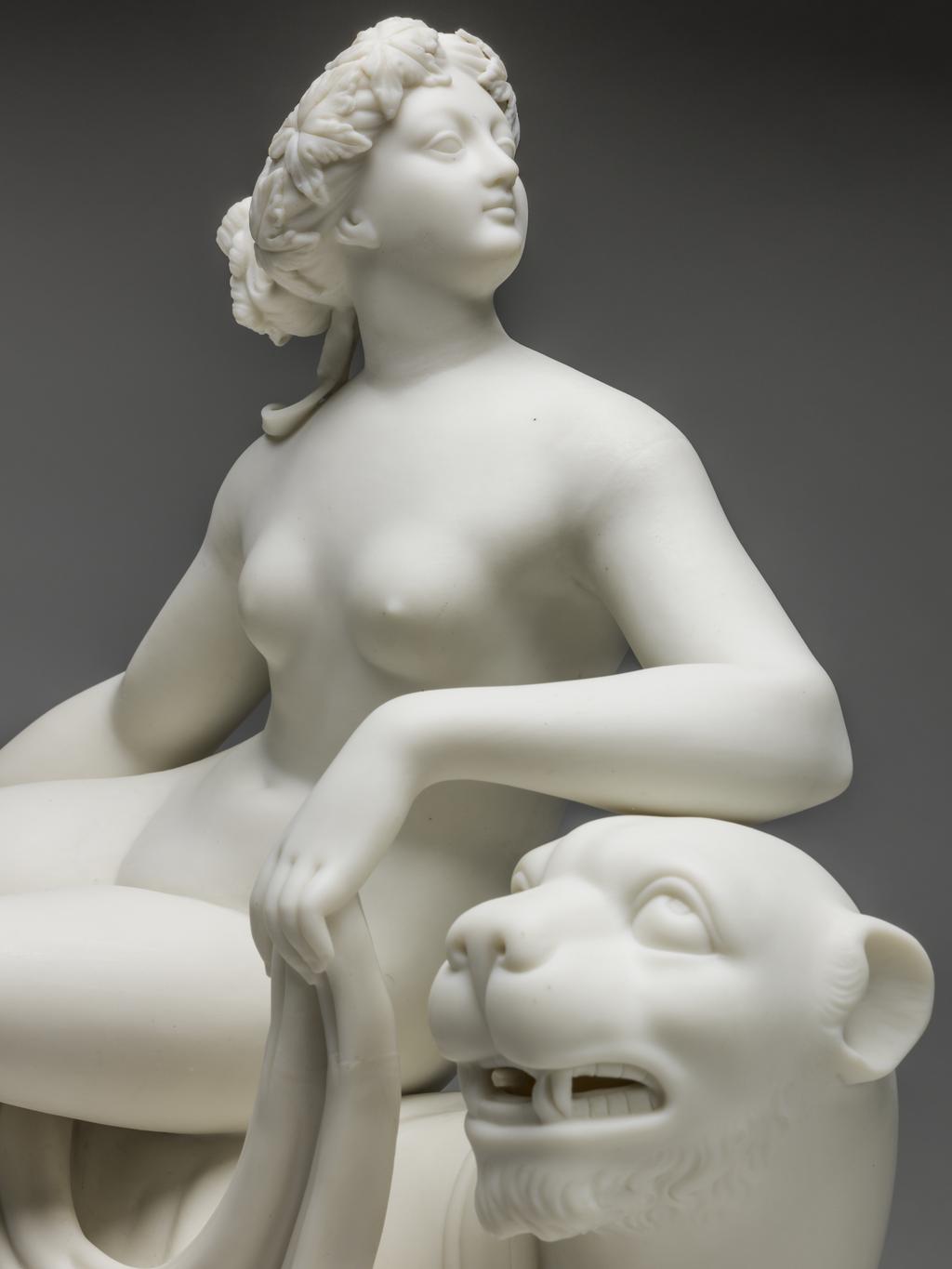 An image of Ariadne on the Panther or Voluptuousness. Minton Factory, Stoke on Trent. After the sculpture by John Heinrich von Dannecker. The figures are supported on a rectangular base with slightly sloping sides and indentations suggesting it is rocky. The panther is walking to the right with its left fore and hind legs forward, its tail between its hind feet, and its head turned towards the viewer, baring its fangs. The nude Ariadne is seated on its back with her right leg extended and her left bent.  Her head is in profile, and she rests her left fore arm on the panther's head. With her left hand she holds up a long drape which extends down to the base at the front, over the panther's back beneath her legs, and down to the back of the base. Creamy-white parian soft-paste porcelain, slip cast and assembled. Height 36.7 cm, length 30.5 cm, depth 14.3 cm, 1852. Neoclassical.