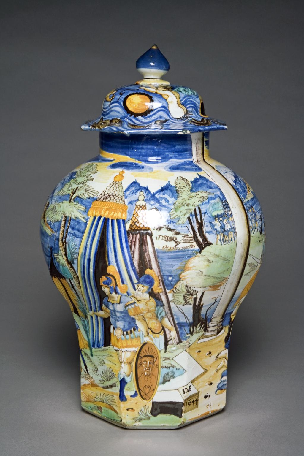 An image of Baluster vase and cover. Conrade Pottery. Lefebvre, Denis. Baluster-shaped with octagonal lower part, and octagonal cover, domed in the centre, and surmounted by a cone-shaped finial which is a replacement in hard-paste porcelain held on by a dowel. The sides are decorated with a continuous scene of Samson and Delilah with in the background, a lake (or the sea), buildings on islands or the shore, and distant mountains. Samson with the ass's jawbone in one hand, lies asleep against the knees of Delilah, who holds a pair of scissors, and has already cut off some of his hair. Behind her are five Phillistines dressed in Roman style armour, one holding a rope to bind Samson. On the right there are two columns and a tent, and on the left, a column and behind it in the distance, a troop of soldiers. On the other side there is a standing soldier on the extreme left, some armour on the ground, and a man to its right pointing towards it. Further to the right are two standing soldiers, one resting his arm on a shield. Behind him are three tents. On the ground to the right of the man wth the shield, there is a stone block inscribed 'DL(joined) F/1644'. The cover is decorated with blue waves on which there is a mermaid holding a torch, a triton, Neptune reclining on a fish with a putto in front of hime, shells, and a large fish. The finial is blue with remnants of gilding. Earthenware, tin-glazed, painted in polychrome, height (whole) 45 cm, diameter (base) 16.5 cm, diameter (rim) 12.4 cm, dated 1644. Nevers, France. Renaissance.