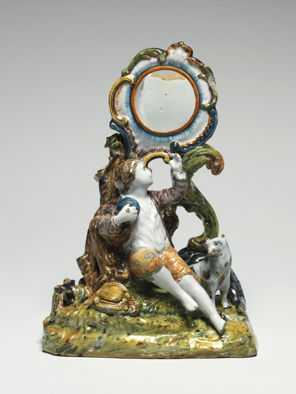 An image of Watch Stand with figures of a Boy and Dog. Unknown, Nièvre, Nevers (formerly in the Nivernais). The rectangular base is open underneath and is decorated on top to resemble a grassy mound. The circular watch holder with a rococo frilled and scrolled edge is supported by a branch of scrollwork and a tree trunk growing upwards from the viewer's left of the base. A barefooted boy holding a blue ball in his right hand and pointing upwards with his left hand sits on the mound with his back to the tree trunk. He wears a white shirt, manganese-purple jacket and pale brown breeches, His yellow broad-brimmed hat trimmed with ribbons lies on the ground to the left. A small white dog with purple and brown patches dog sits to the right of him. Earthenware, moulded in parts, tin-glazed, and painted in blue, olive-green, yellow, manganese-purple, purple, pale yellowish-brown and dark brown high-temperature colours, height, whole, 24.5 cm, width, whole, 17.5 cm, circa 1760. Rococo.