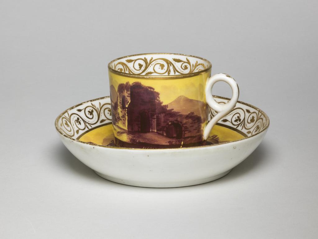 An image of Coffee can and saucer/cup and saucer. Minton factory, Staffordshire, Stoke-on-Trent. The can is cylindrical with a circular ring handle. The saucer has deep curved sides and stands on a footrim. The cup is decorated externally with a yellow ground on which is painted a dark maroon landscape with a ruin on the right, and a low farm house and a tree on the left with mountains in the distance. There is a gold band below and another on the rim. Inside at the top there is a border of gold foliated scrolls. The cup is decorated en suite with ruins in a landscape with trees and distant mountains radiating from two gold concentric circles in the middle. Below the the rim there is a border of foliated scrolls between gold horizontal bands. Pattern no. 604. Bone china (?) decorated with a bright yellow ground, painting in dark maroon enamel, and gilding, height, cup, 6 cm, diameter, cup, 6.8 cm, width, cup, 8.3 cm, height, saucer, 3.2 cm, diameter, saucer, 13.8 cm, circa 1800. Production Note: The decoration imitates French late 18th porcelain, decorated with yellow grounds and monochrome purple or dark puce landscapes. On Minton, the painting is often executed rather roughly.