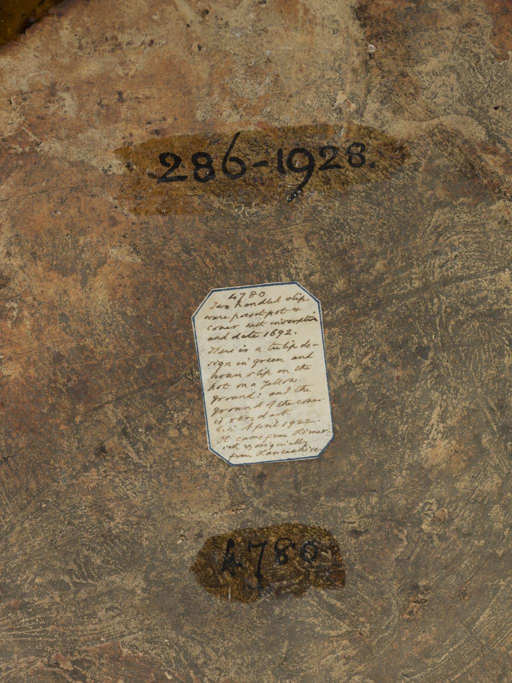 An image of Posset Pot/Two-handled pot and cover. Staffordshire Pottery, unknown maker. Red earthenware, thrown, coated inside and out with white slip, and slip-trailed in dark red, olive-green, and white slips under yellowish lead glaze. Cylindrical with two S-shaped handles. Decorated round the top with a slip-trailed inscription, 'THE BEST IS NOT TOO GOOD FOR YOV 1692'. Below this, on each side, is a pair of stylized tulips separated by vesica-motifs, the latter made up of studs of slip with star impressions of a cut stick. The lid is left red without a coating of white, and is decorated with quatrefoils in white ouline filled in with olive-green slip showing the impression of a textile mesh. 1692. William III and Mary II. No dimensions recorded. Production Note: The cover is almost certainly a 'marriage'.