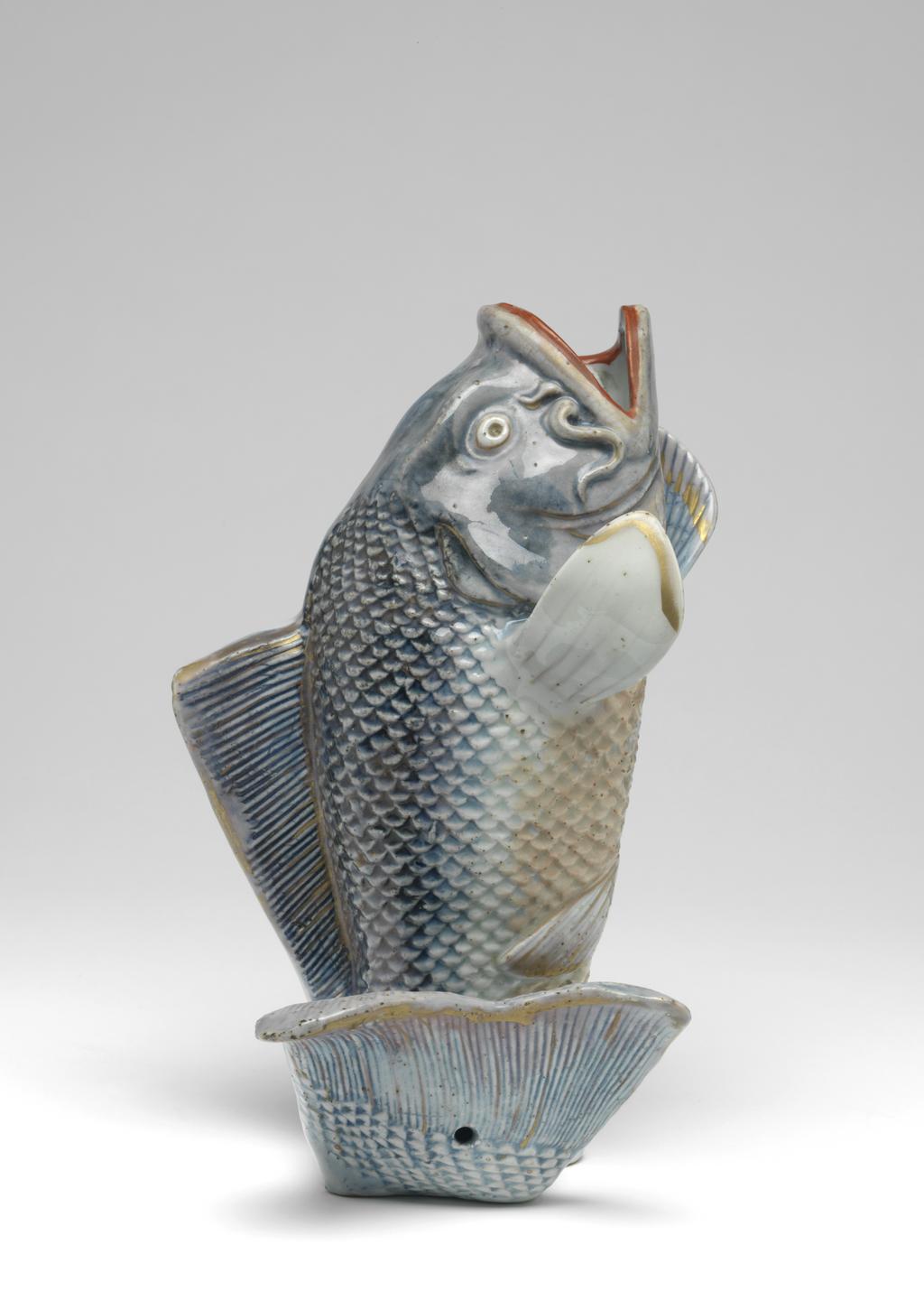 An image of Carp-shaped wall vase. Production Place: Japan, Arita. The carp, hollow-moulded, leaps upwards supported by its curling tail and rests on a simple base with an uneven and gritty underside. The body is finely moulded with scales and gills, and the fins and tail finely incised. The head, upper half of the body, dorsal fin and tail are covered in a wash of underglaze blue, the lower half with pale red enamel and covered with a bluish glaze; details of the tail, fins and head are outlined in gold; the large open mouth is picked out in red and gold and a beady eye in gold with a recessed pupil. The back of the model is flat and undecorated except where the underglaze blue has flowed. A hole is pierced in the centre of the back for suspension and at the base of the tail. Hard-paste porcelain decorated underglaze in blue and overglaze in red, pale-red, gold and violet enamels, and gold, height, whole, 24.2 cm, diameter, whole, 13.3 cm, width, whole, 15.5 cm, circa 1700-1800.