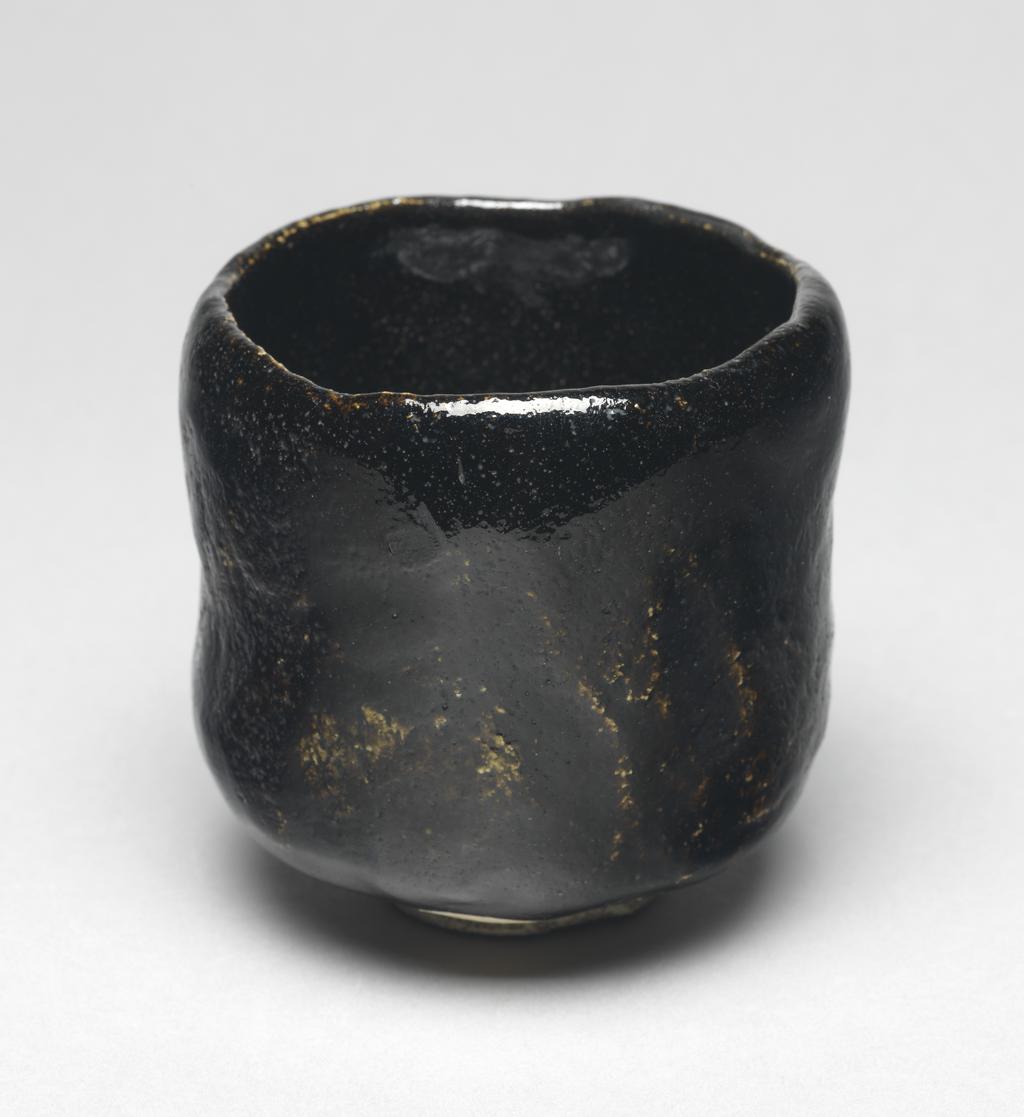 An image of Japanese pottery. Tea bowl. Ryonyu (Raku IX). Production Place: Japan, Kyoto. Hand-built footed tea bowl of very coarse grey-white earthenware with a thick black Raku glaze. Raku spiral in foot ring. Hollow at the centre of well. Vertical slicing on one side. Earthenware, glazed, height 9.40 cm, diameter, bowl, 10.00 cm, diameter, foot, 4.40 cm, circa 1775-circa 1825. Edo Period (1615-1868).