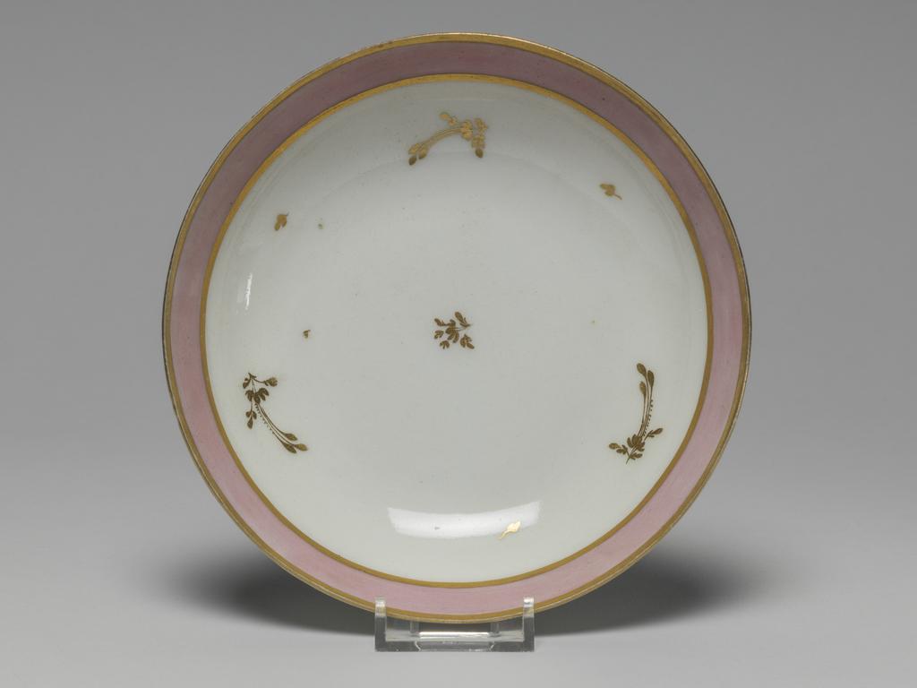 An image of Saucer. New Hall Porcelain Factory, Staffordshire. Hybrid hard-paste porcelain saucer, from a teaset. Decorated overglaze with a wide band of pink enamel between narrow gold bands and with scattered floral sprigs in gold. Pattern number 223. Height 3.2 cm, diameter 13.1 cm, 1785. See C.11A-L-1988: A service comprising a teapot and cover of 'silver' shape, a milk jug, sugar basin and cover, a slop basin, two plates of different sizes, two tea bowls, two coffee cups and two saucers.