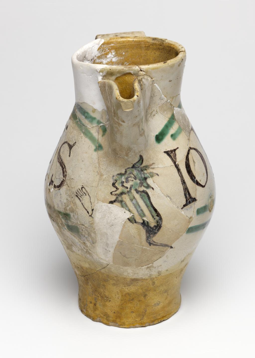 An image of Maiolica arcaica. Jug. Unidentified Orvieto pottery, Italy, Umbria. Shape 7. Ovoid with flat base; cylindrical neck, expanding slightly towards the rim; broad strap handle; spout made from a strip of clay, folded and pressed to the neck. On the front under the spout, a grotesque devil with a striped body, long tail and four horns on its head, faces to the left holding out one of its hands. One side of the jug bears the word `SaS' (Sanctus) in manganese, enclosed by two green lines forming a curved rectangular frame, open on the side nearest the devil. The other side bears the word `IohS' (Johannes), similarly framed. Cream earthenware, the lower part and interior are lead-glazed brownish-yellow, the rest is tin-glazed off-white; the tin-glaze has crazed; painted in manganese and copper-green. Height, whole, 22.7 cm, diameter, base, 8.9 cm, diameter, body, 14.3 cm, width, whole, 17.6 cm, circa 1300-1350. Medieval.