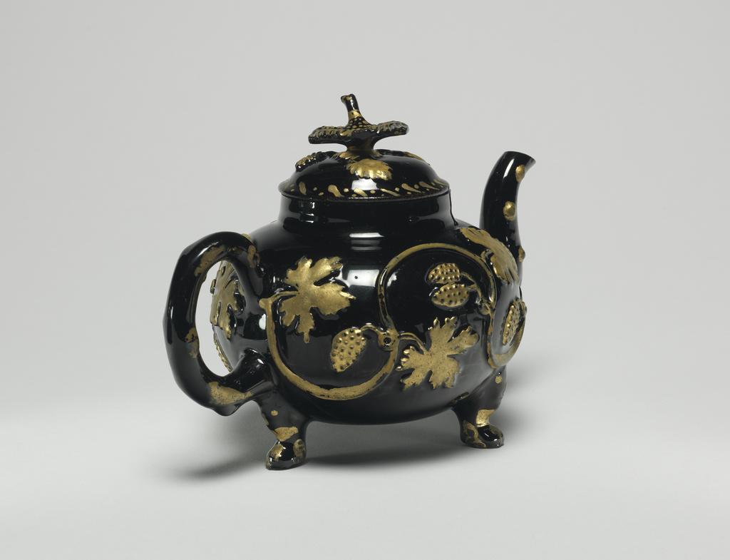 An image of Teapot and cover. Unidentified Staffordshire Factory. Dark red earthenware, the body thrown, with applied moulded handle, spout, feet, and finial, and applied moulded sprigs, the whole covered with black lead-glaze, and partly gilded. The pot has a depressed globular body supported on three lion's mask and paw feet, a crabstock handle and spout, and a domed cover with a bird finial. Both sides of the body are decorated with a coiling vine-stem with bunches of grapes and foliage in relief, which have been oil gilded. Rococo. No dimensions on Adlib (07/2014).