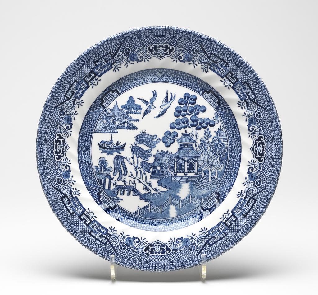 An image of Willow Pattern Plate. Wessex Collection. Circular with a slightly wavy edge, sloping rim with slanting fluting, shallow sides and flat centre, with a raised ring on the reverse. The flat area is occupied by the Willow Pattern with three figures on the bridge and two bvirds flying overhead. This is surrounded by a border comprising four wide V shapes enclosing a rectangular motif with incut corners, alternating with panels of ribbon diaper. The rim has a broad dense border comprising panels, plants, rectangular and circular motifs, with a small diaper patterned background, and round the edge, radiating rows of tiny circles. White ceramic body, moulded, and printed underglaze in blue. Height 2.5 cm, diameter 25.5 cm, circa 2000-2015. Chinoiserie.