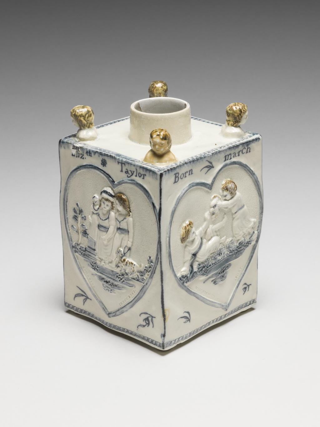 An image of Pearlware Tea Caddy. Bovey Heathfield Pottery, Devonshire. On each corner there is an applied moulded bust of a child, and on the base four coiled feet. Two opposing sides are decorated with a heart-shaped raised outline enclosing a relief of a little girl holding a doll accompanied by a little boy with a small dog on a lead, with a small tree and a low broken off colum on the left. Below them is an impressed inscription, 'SPORTIVE INNOCCENCE' .On the other two sides there is a similar heart enclosing a relief of a child holding out a bearded male mask to frighten a little girl who has fallen to the ground and raises her left hand to push it away. Below is the impressed inscription, 'MISCHIEVOUS SPORT'. In each of the lower spaces outside the heart outlines there is a painted sprig, and above one, a moth or butterfly. Painted round the top of three sides is the inscription, Eliz.th (th raised) Taylor/Born march/18th. (th raised) 1795'. Cream earthenware, moulded. Pearlware, painted underglaze in dark blue and yellowish-brown, dated 1795. English.