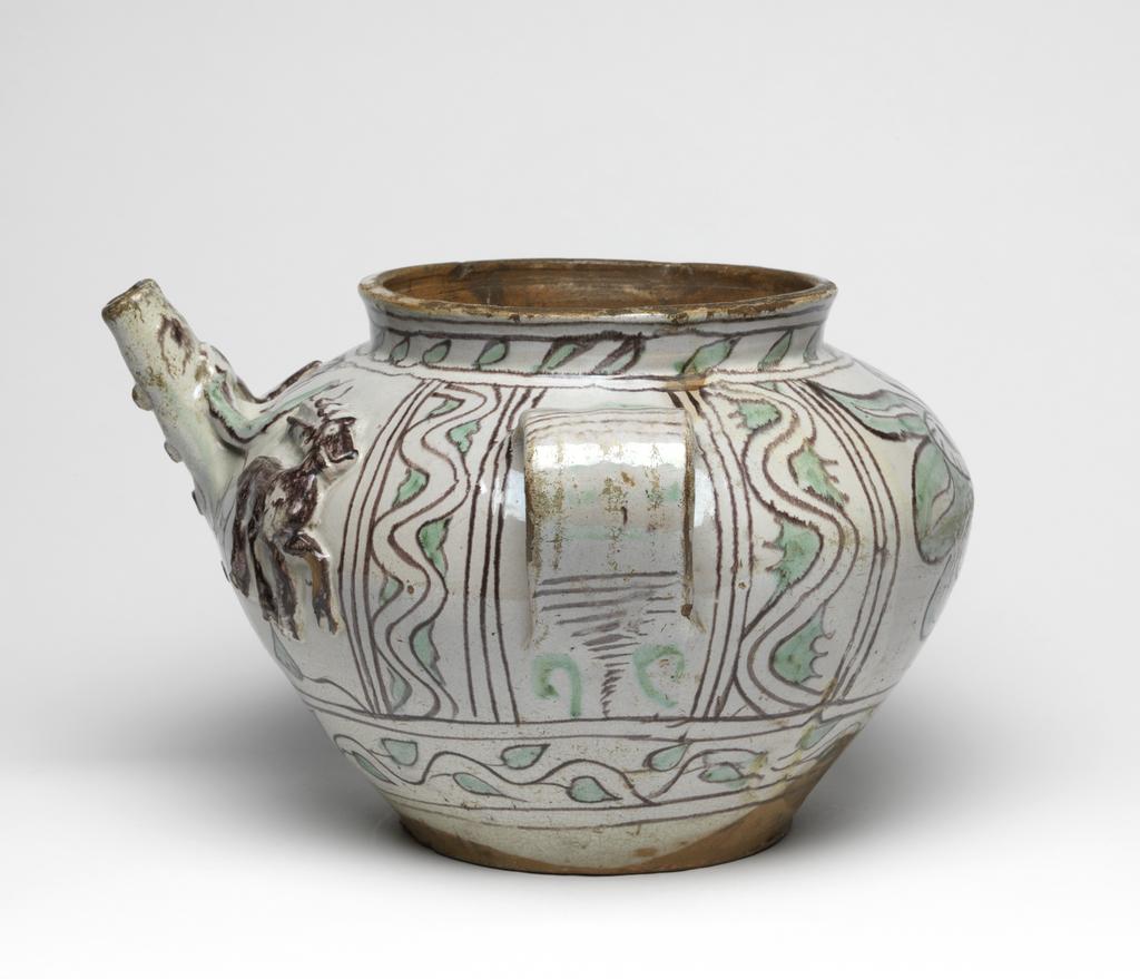 An image of Maiolica. Two-handled spouted jar. Unknown, Tuscany, Umbria or Lazio. Bulbous body narrowing sharply towards the flat base, with short cylindrical neck and two broad strap handles. At centre front there is a straight, upward-sloping spout with horns above, four spots below, and a stag on each side, applied in relief. The spout is decorated to resemble a buffalo's head. The other side is decorated with a stylised flower with two peacock's tail eye motifs in its centre. The handles are flanked by two sets of three vertical manganese lines enclosing three wavy stems with leaves at intervals. The lower part is encircled by an undulating spray of leaves between horizontal manganese bands, and the neck by slanting spaced leaves between pairs of manganese horizontal bands. On the handles, there are alternate groups of manganese and green horizontal lines, flanked, at the bottom, by two addorsed C-shaped strokes. Pale red earthenware, the interior lead-glazed brownish-yellow, the exterior tin-glazed greyish-white; base unglazed. Painted in manganese and pale green. Height, whole, 20.3 cm, diameter, base, 14.3 cm, width, whole, 31.8 cm, circa 1400-1450. Late Medieval