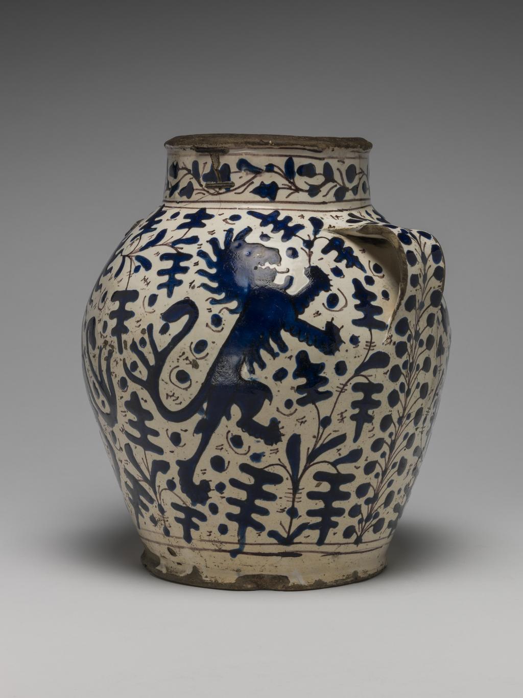 An image of Maiolica. Pharmacy or storage jar. Unknown maker, Florence. Both sides are decorated with two lions rampant addorsed, each holding a spray of oak leaves in its raised paw, surrounded by sprays of oak leaves with dots in the background. Round the lower part of the body there are two horizontal manganese bands. On the neck, between pairs of horizontal manganese bands, there are oak leaves on a wavy stem. On each handle, and continuing down the side of the jar beneath it, is a fern frond or stalk with twigs branching off on each side and terminating in small round berries. Buff earthenware, tin-glazed creamy-white on the interior and exterior; the rim and base unglazed. Painted in bright, extremely shiny relief-blue with manganese outlines. Height, whole, 25.8 cm, diameter, mouth, 12.4 cm, diameter, base, 14.5 cm, width, across handles, 23.0 cm, circa 1420-1450. Renaissance.