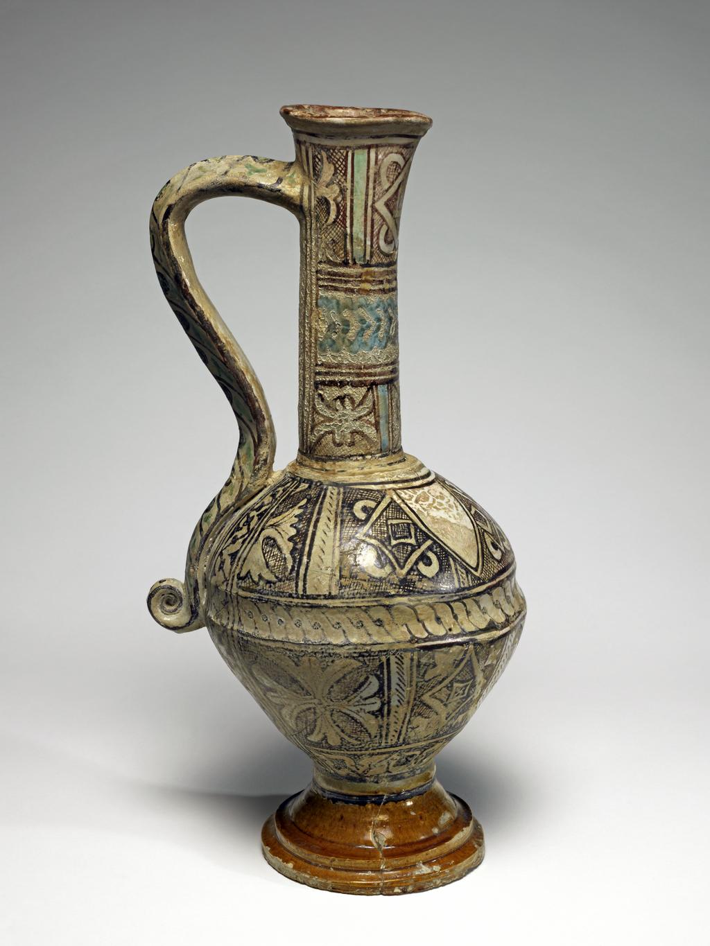 An image of Tin-glazed earthenware ewer. Maiolica arcaica. Late Medieval, painted in copper-green, turquoise-green and manganese with panels containing stylised foliage, rhomboids and knots, surrounded by cross-hatching and shields. Height 38.0cm, diameter, foot, 11.5cm, width, 19.0cm. Orvieto, Umbria. Circa 1275-1350.