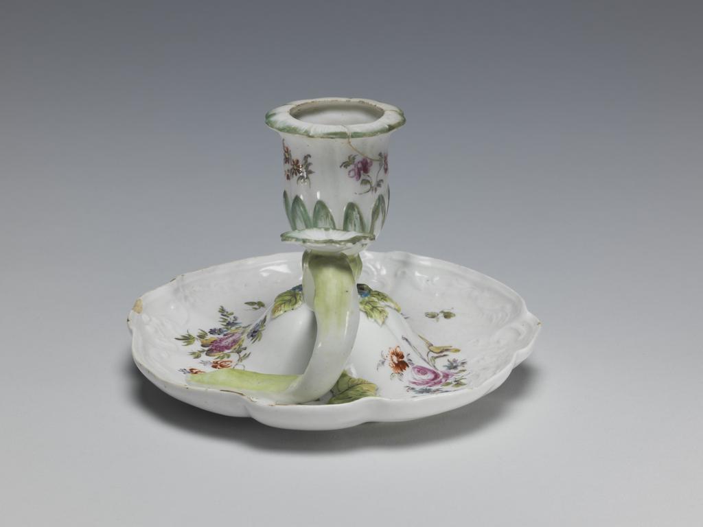 An image of Chamber Candlestick/Hand-Candlestick. Derby Porcelain Factory. The circular dish has curved sides with four large and four small lobes round the edge and a border of raised scrolls and foliage. It rises up in the centre to support the inner end of the handle and the nozzle in the form of a bell-shaped flower with a calyx of pointed leaves. The handle is in the form of a branch which curves upwards and downwards to rest on the dish. At its hightest point it has a horizontal flower-shaped thumbpiece. On the dish around the junction with the nozzle there are three blue flowers each with a pair of leaves, and another leaf lies further out beside the lower end of the handle. The dish is painted with three large sprays of mixed flowers and the nozzle with three smaller sprays of flowers of tone type. The outer edge has remnants of gilding. Soft-paste porcelain candlestick, moulded, lead-glazed, and decorated with applied flowers and leaves, and painting in polychrome enamels, height, whole, 8.2 cm, width, whole, 12.5 cm, circa 1760.