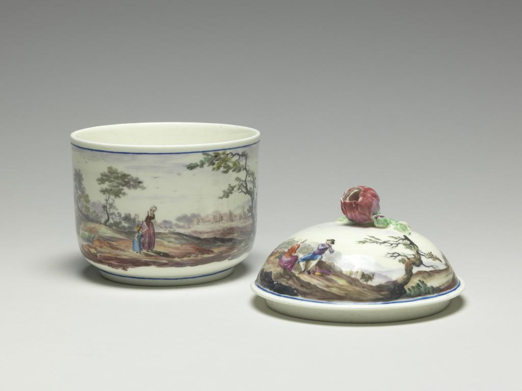 An image of Teaware. Sugar basin and cover. Mennecy Porcelain Manufactory. The cover is domed and has a knob in the shape of a half open rose bud, with a small rosebud and three leaves on a twig resting on the cover. The bowl is decorated with a continuous landscape in which on one side there is a distant village and a woman and a child, and on the other a lady and two gentlemen seated on rocks beside a lake. Above and below there is a narrow blue horizontal band. On the cover there is a different landscape in which there are two sheep on one side, and on the other, a goatherd playing a pipe to a seated woman, and a goat in the background to right. A narrow blue band encircles the edge of the scene. The knob is pink with pale green leaves. Soft-paste porcelain, the bowl probably thrown, lead-glazed, and painted in blue, green, greyish-green, dark pink, red, purple, several shades of brown, grey and black enamels. Height, overall, 12.4 cm, height, bowl, 7.3 cm, diameter, bowl, 9.7 cm, diameter, cover, 10.2 cm, circa 1760. Rococo. Louis XV.
